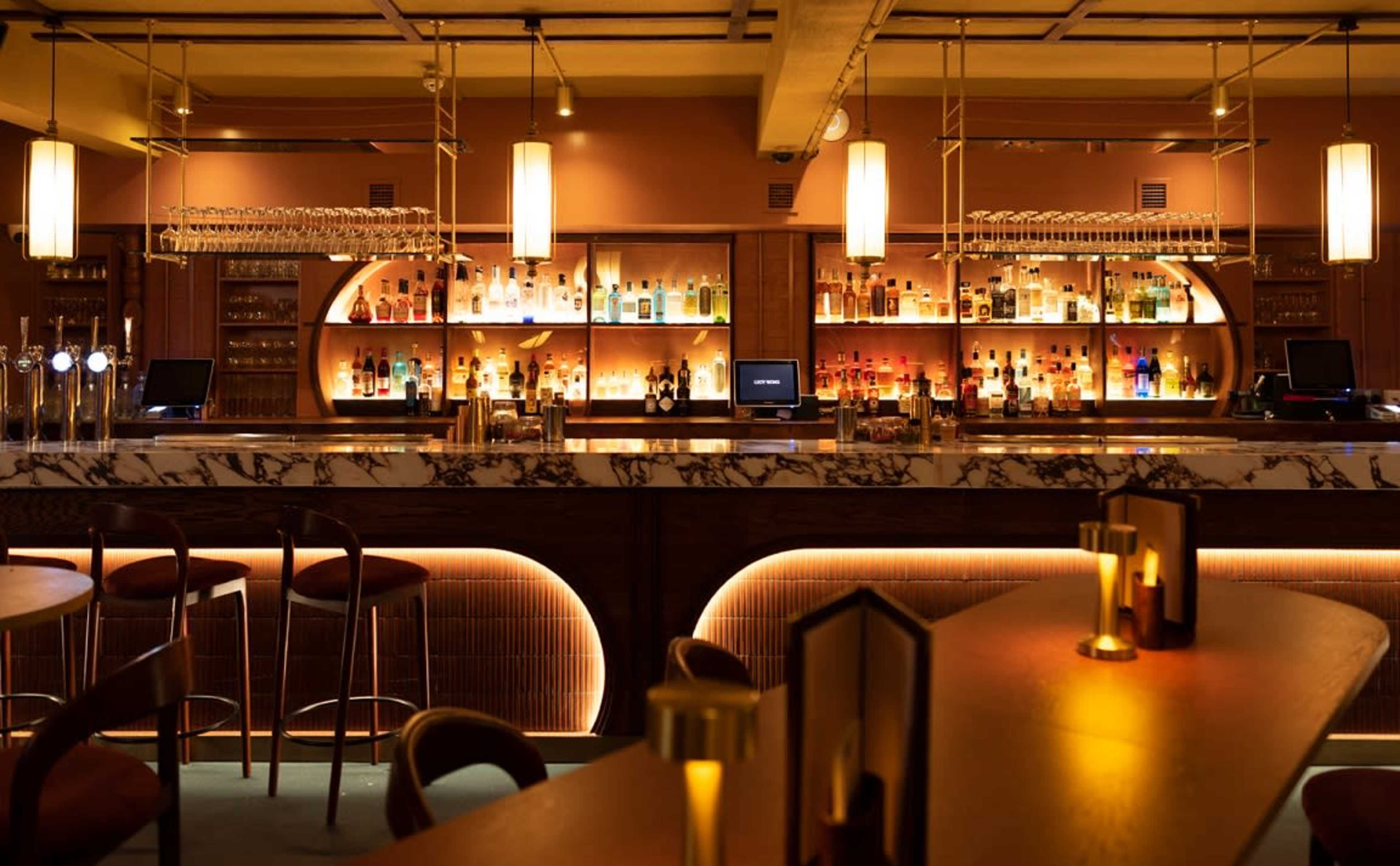 The image shows a dimly lit bar with a marble countertop, illuminated shelves of liquor bottles, and several wooden stools arranged around it.
