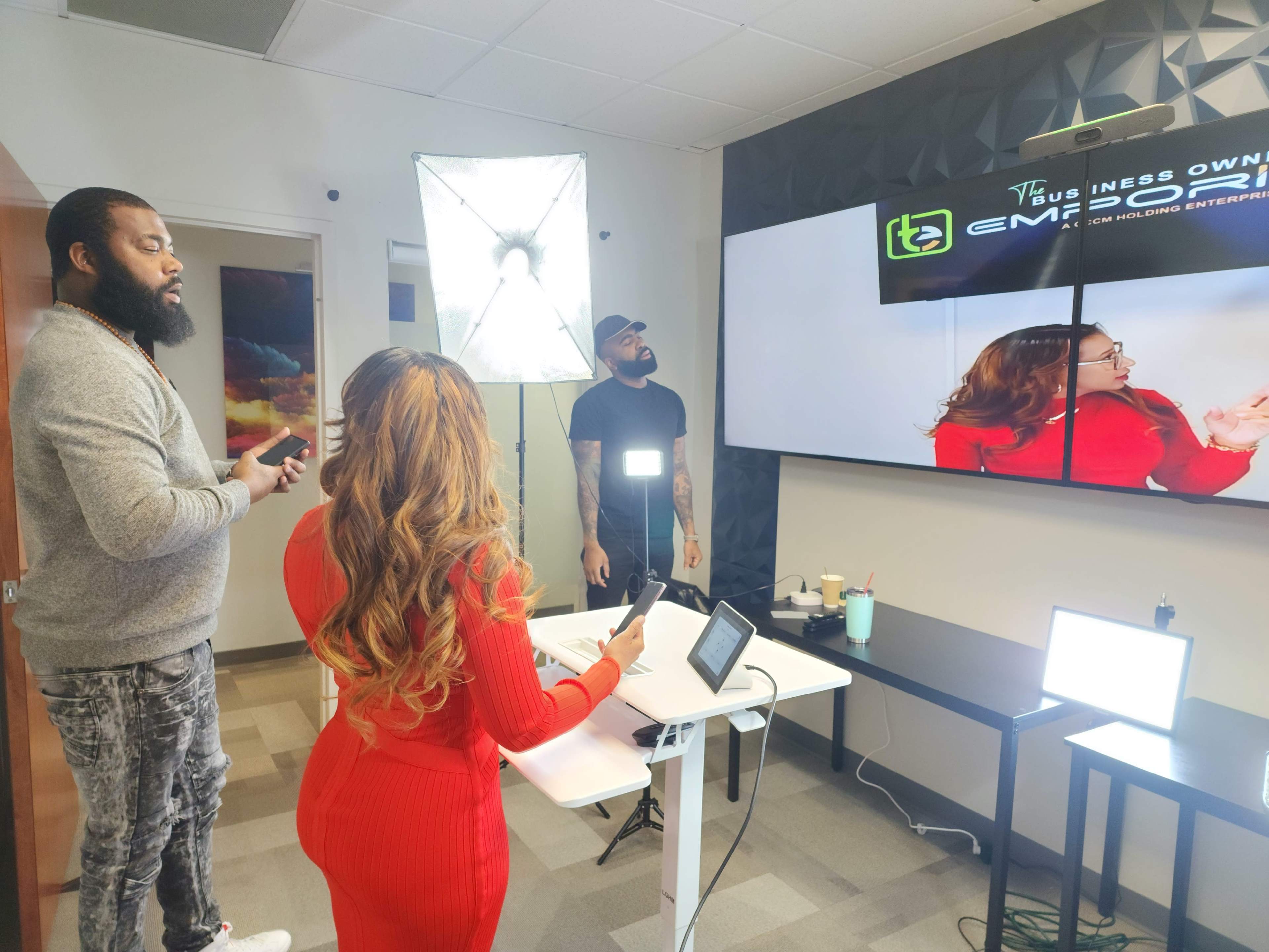 A woman in a red dress stands at a table, looking at a screen while two men observe, one holding a camera and the other adjusting lighting equipment in a modern office setting.