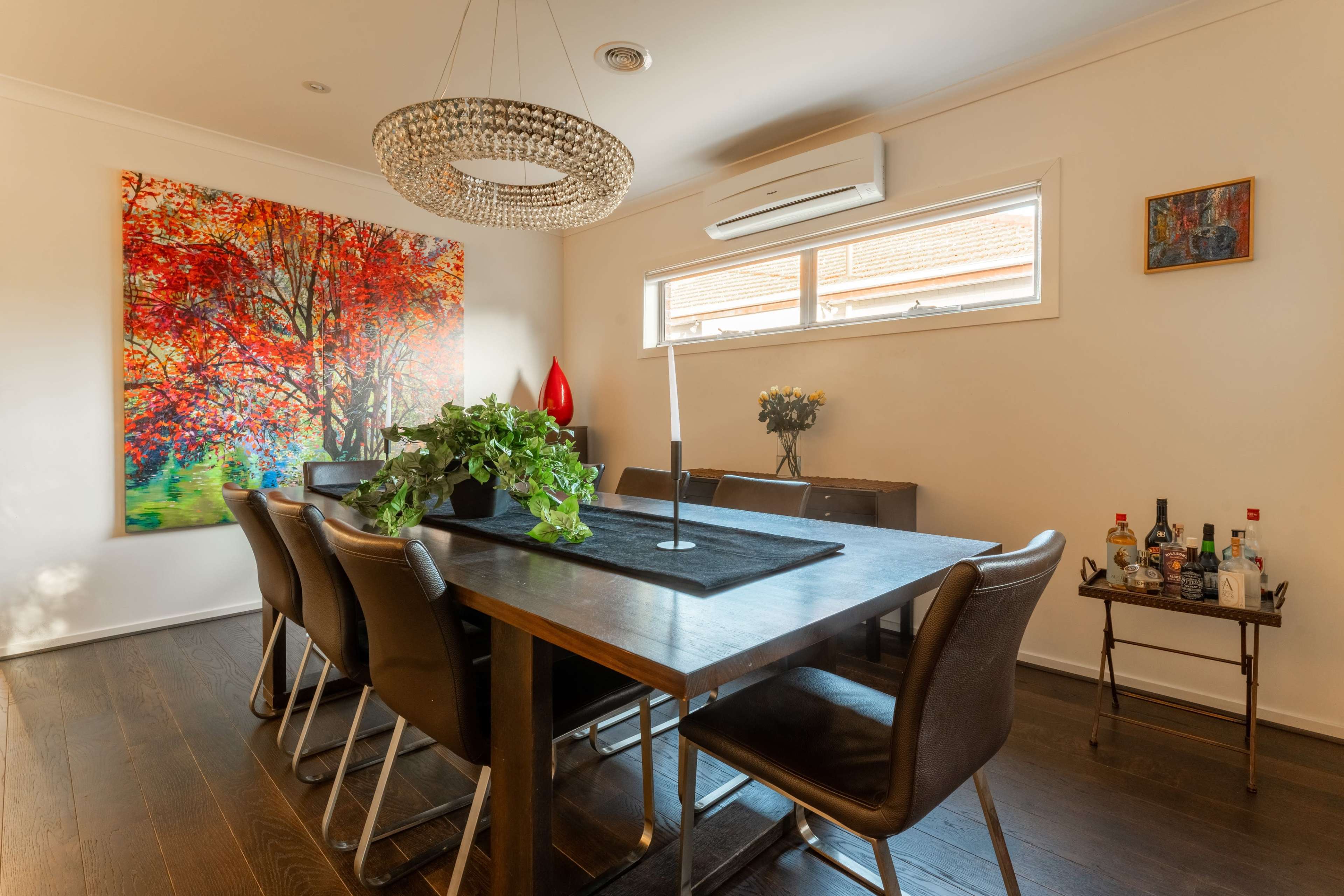 A dining area features a large wooden table surrounded by black leather chairs, with a vibrant painting on the wall and a drinks cart in the corner.