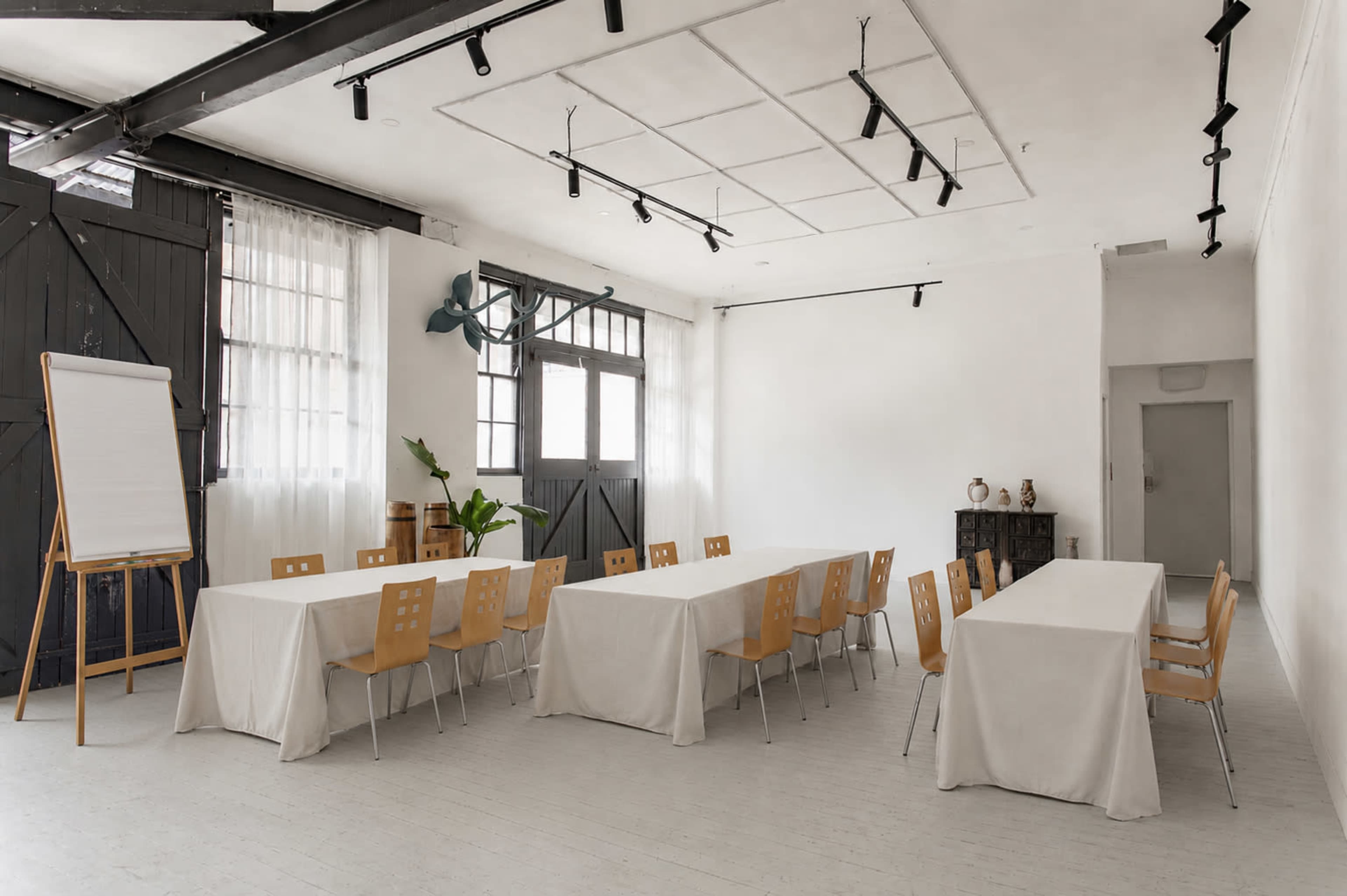 The image shows a spacious, well-lit room set up with several long tables covered in white tablecloths and surrounded by wooden chairs, along with a flip chart and a plant in the corner.