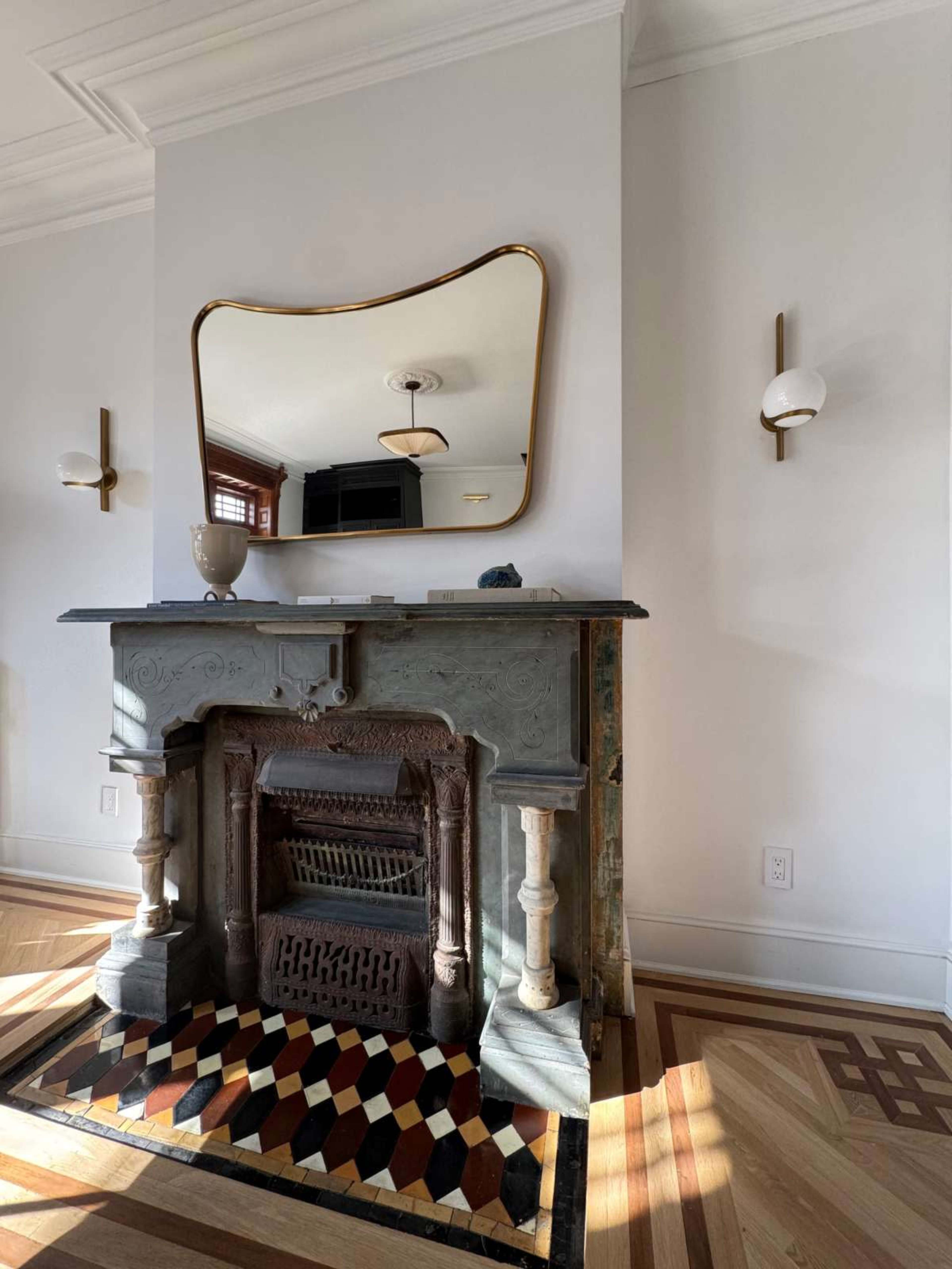 The image shows a decorative fireplace with an ornate mantel, a large mirror above it, and wall sconces on either side, set in a well-lit room with patterned flooring.