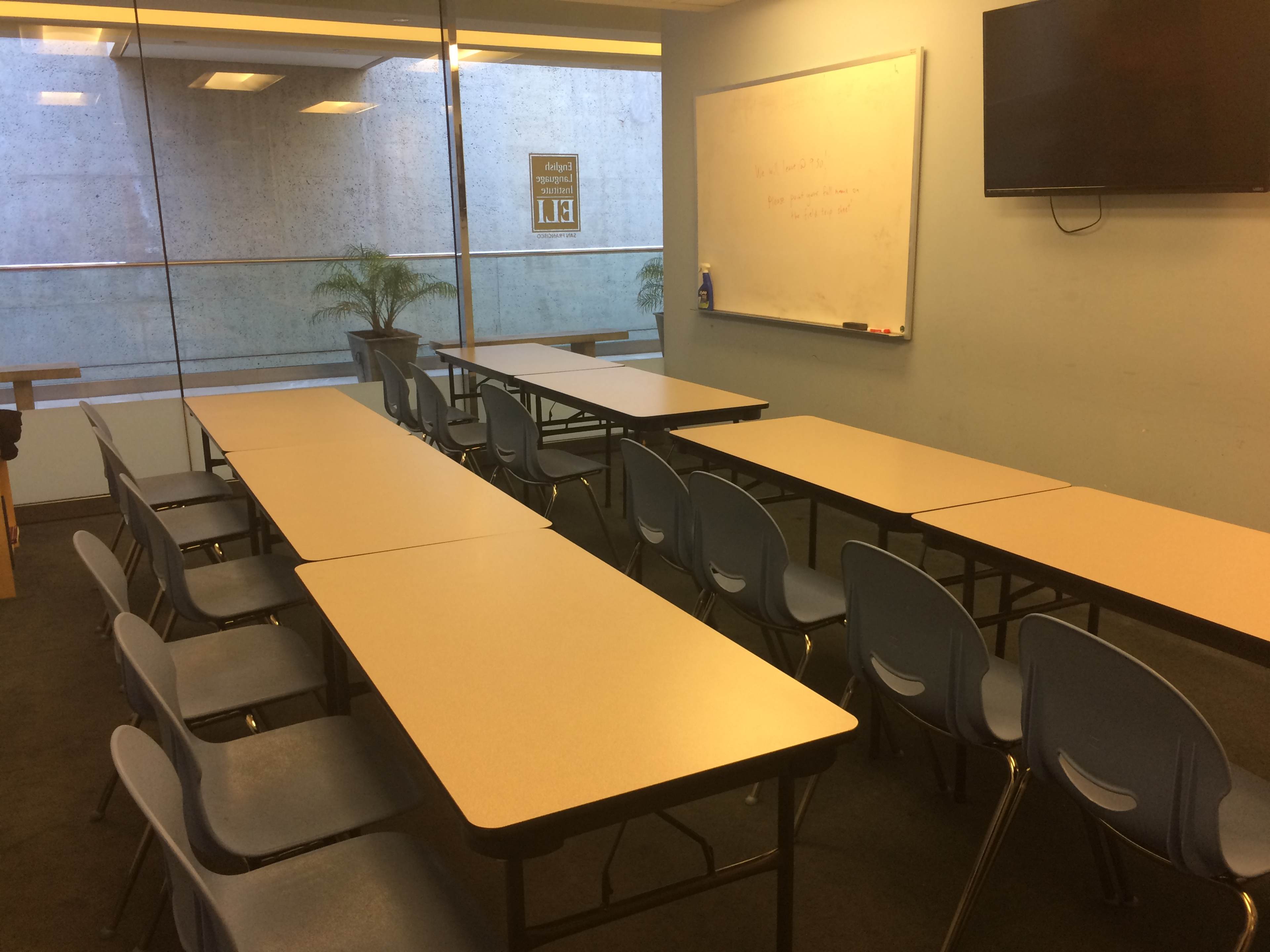 The image shows a classroom with several tables arranged in two rows, each accompanied by blue chairs, and a whiteboard and TV mounted on the wall.