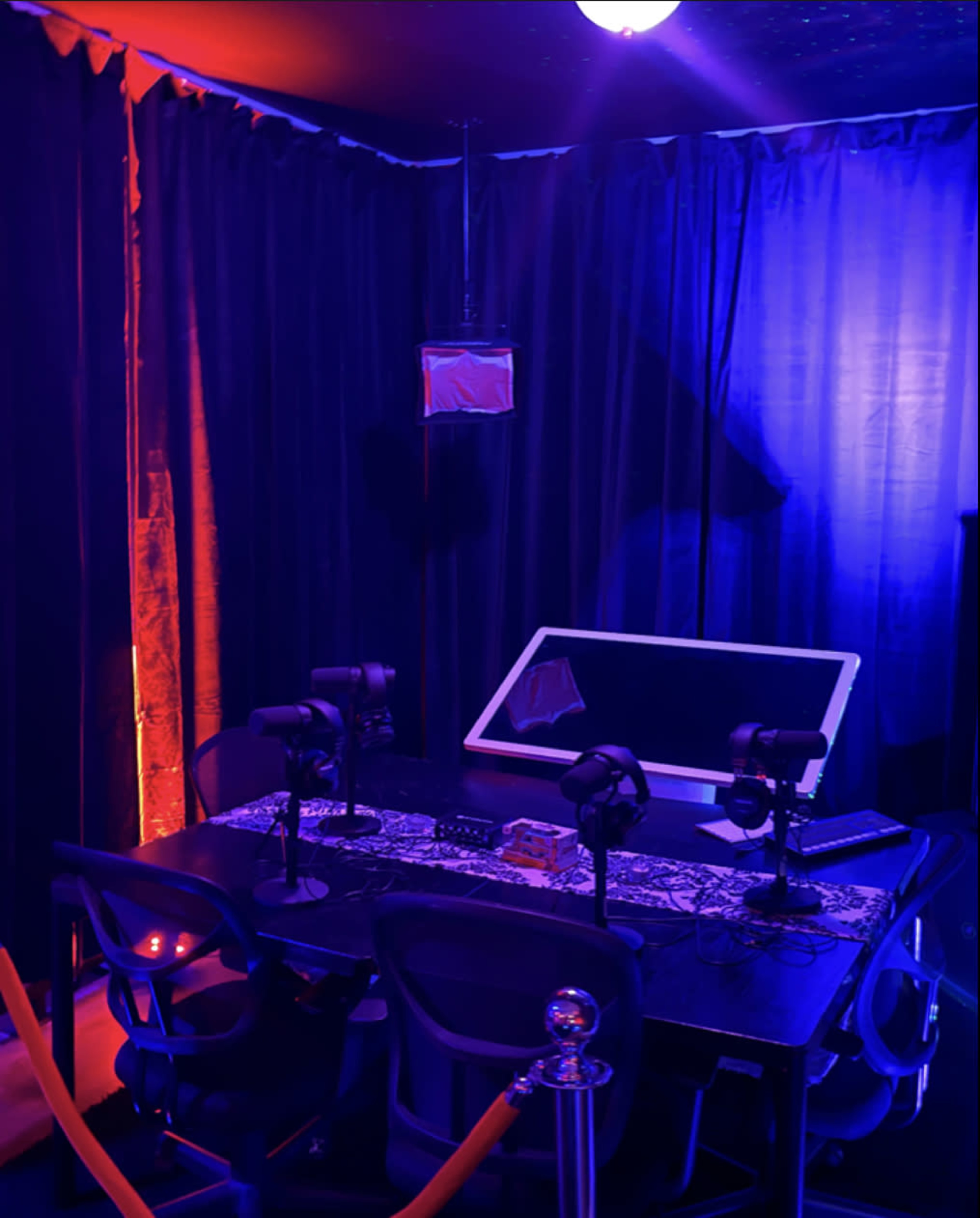 A dimly lit room with blue and purple lighting, featuring a table with microphones and a large monitor surrounded by dark curtains.