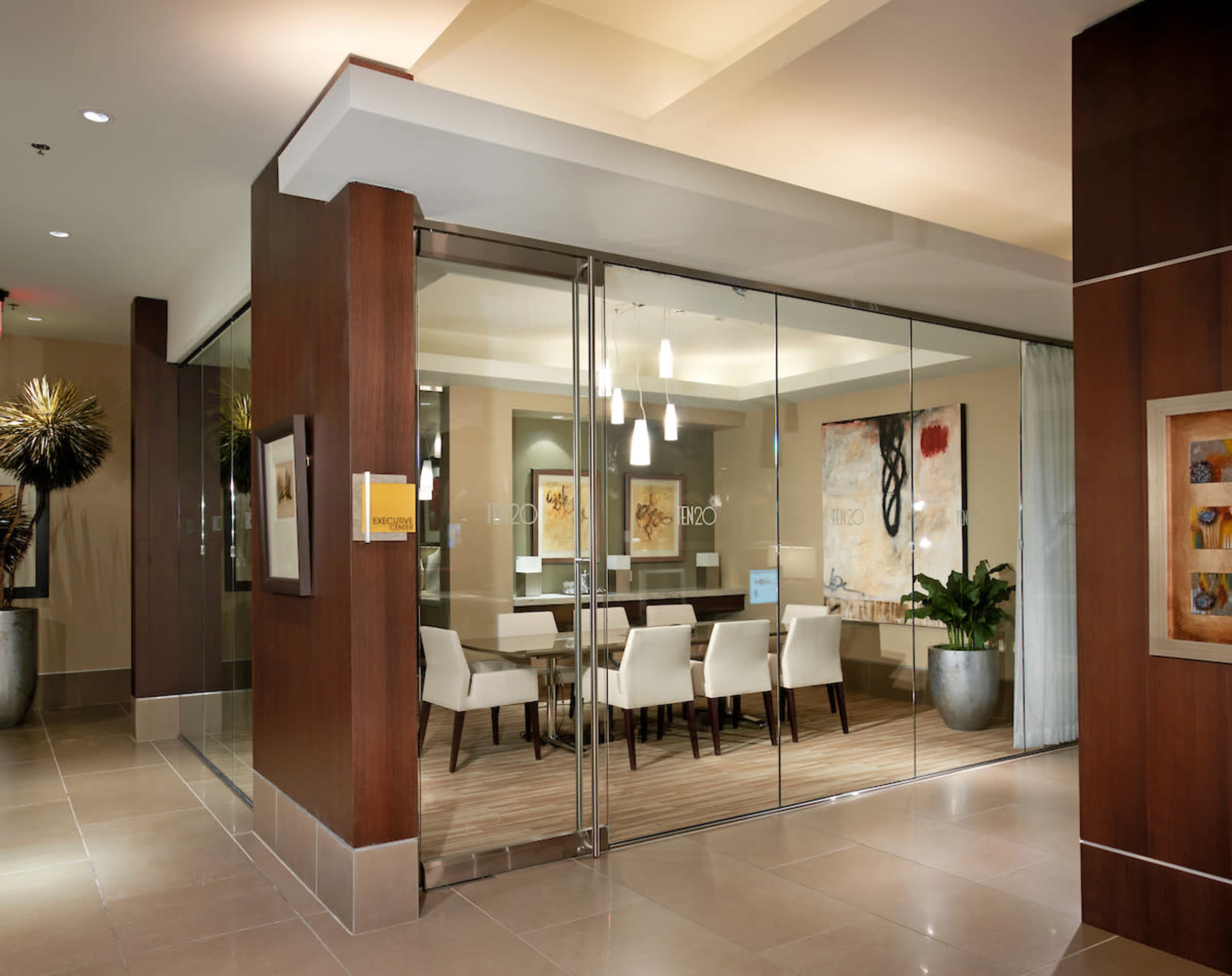 A modern conference room with glass walls, featuring a wood-paneled entrance, white chairs around a large table, and decorative artwork on the walls.