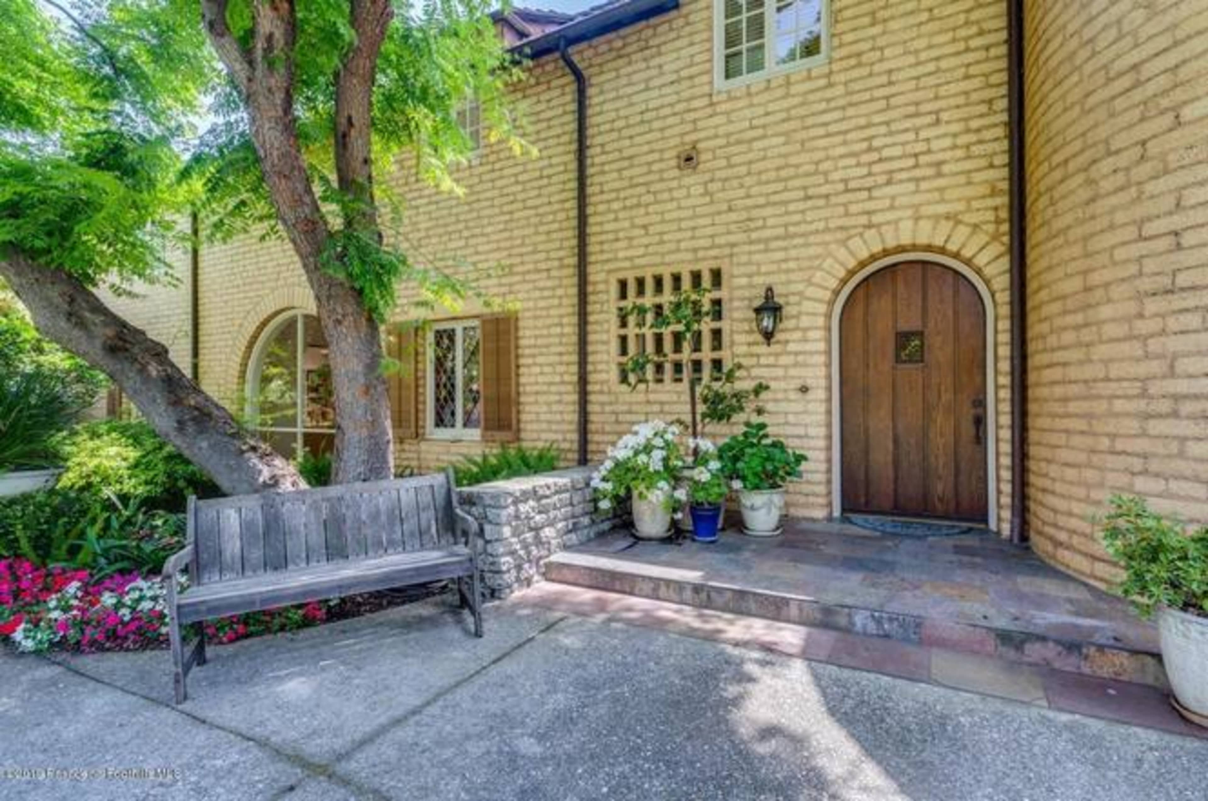 The image shows a brick house entrance with a wooden door, a stone pathway, a wooden bench, and landscaped flowerbeds.