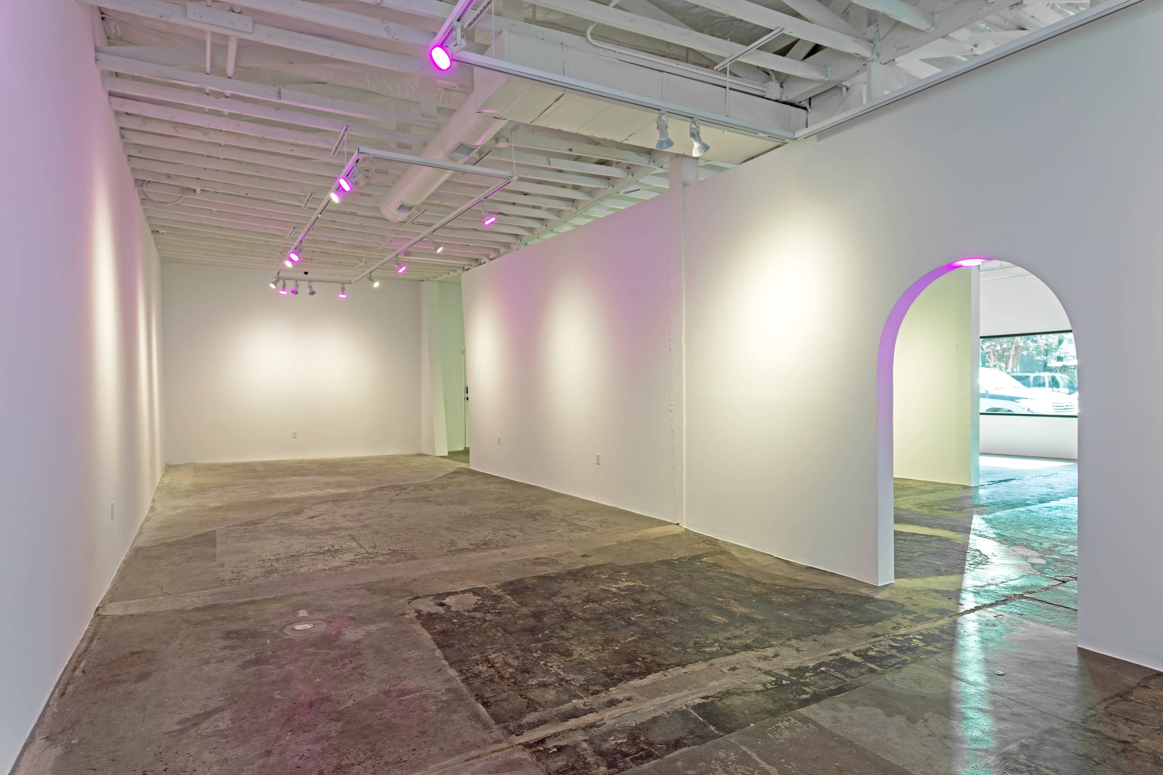 A minimalist art gallery with a sparse layout, featuring concrete flooring and white walls, illuminated by pink lights.