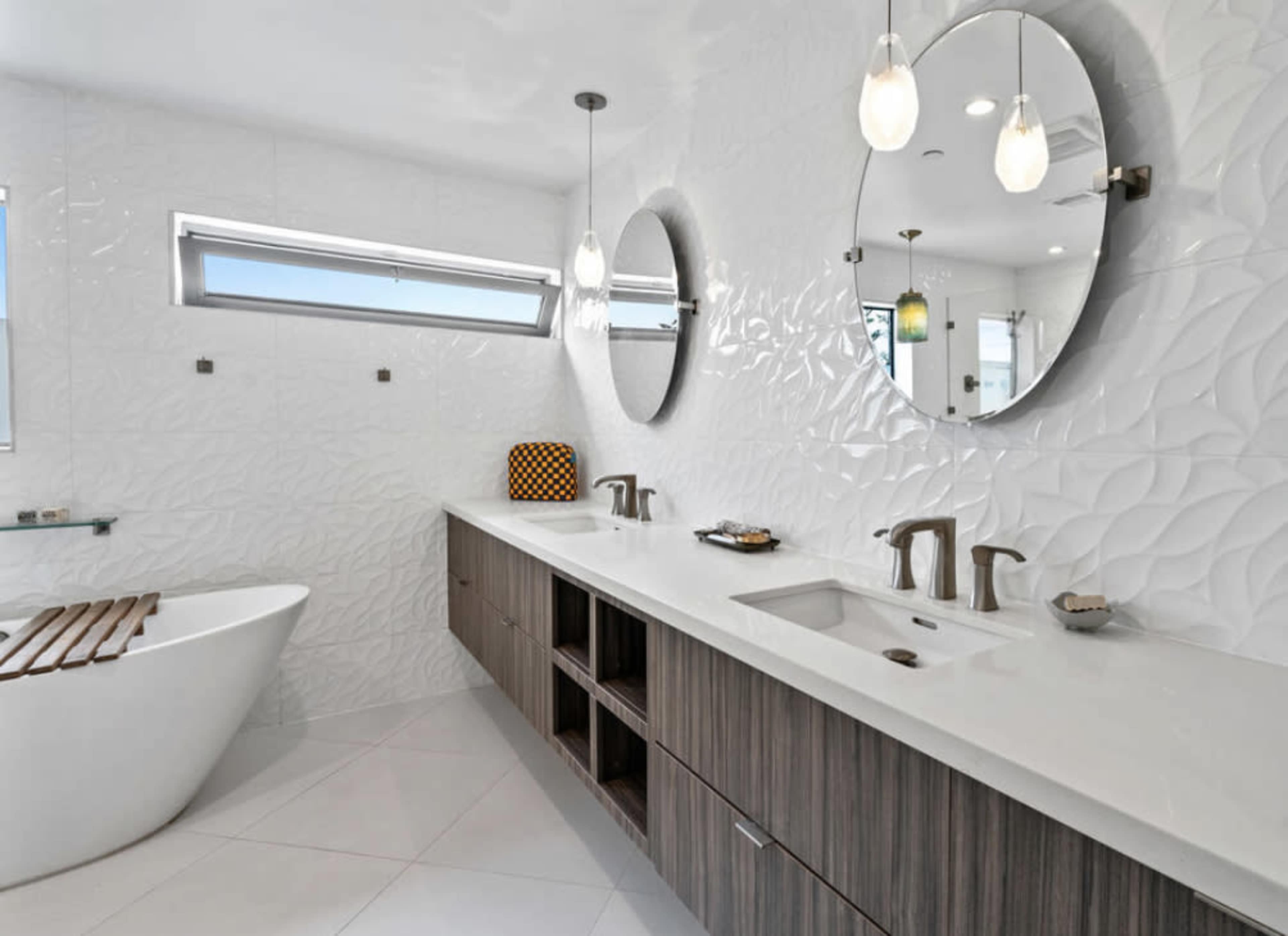 The bathroom features a freestanding tub, double sinks with a wooden vanity, and two round mirrors above the sinks, all accented by textured white walls and ample natural light.