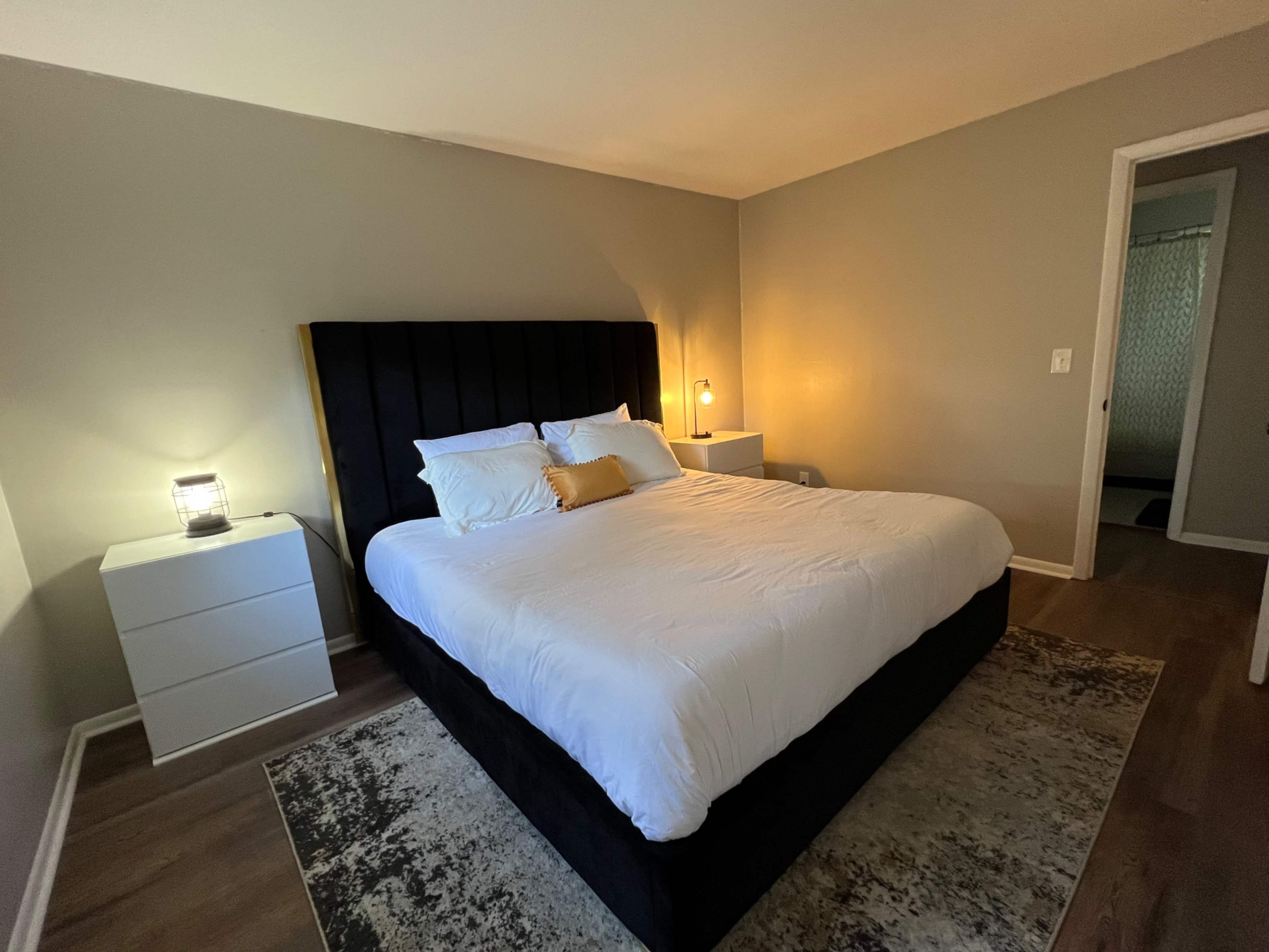 A modern bedroom with a large bed, a nightstand with a lamp, and a doorway leading to another room.