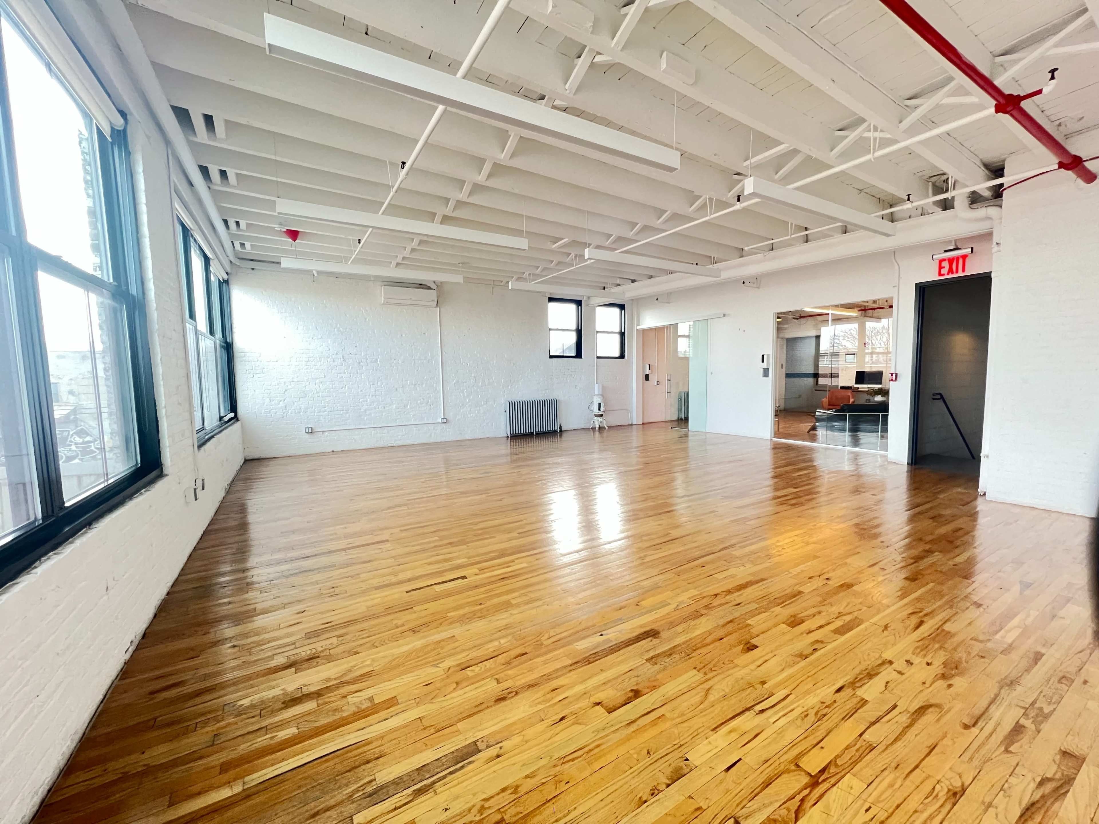 A spacious, empty room with polished wooden floors and large windows, featuring exposed ceiling beams and an exit door.
