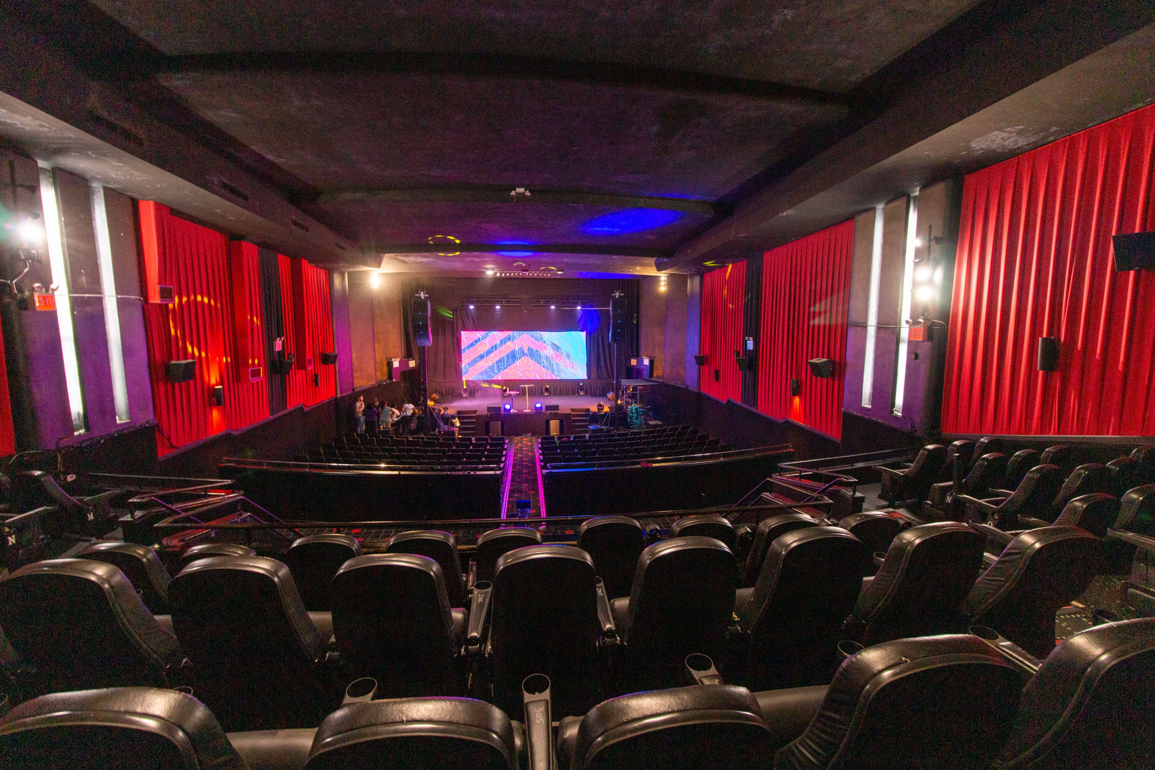 A view from the back of a theater featuring rows of empty black seats facing a stage with a colorful backdrop and red curtains.