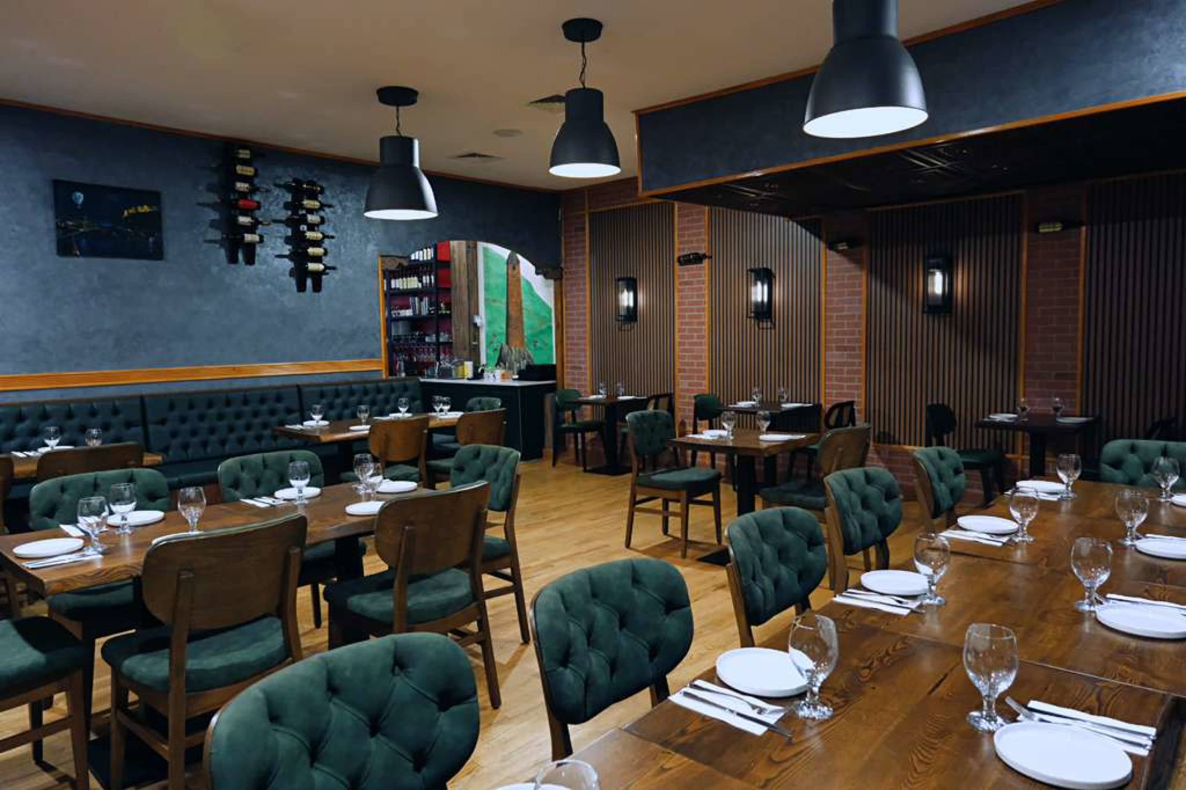 The interior of a restaurant features wooden tables arranged with plates and glasses, surrounded by green upholstered chairs and walls with a mix of textures.