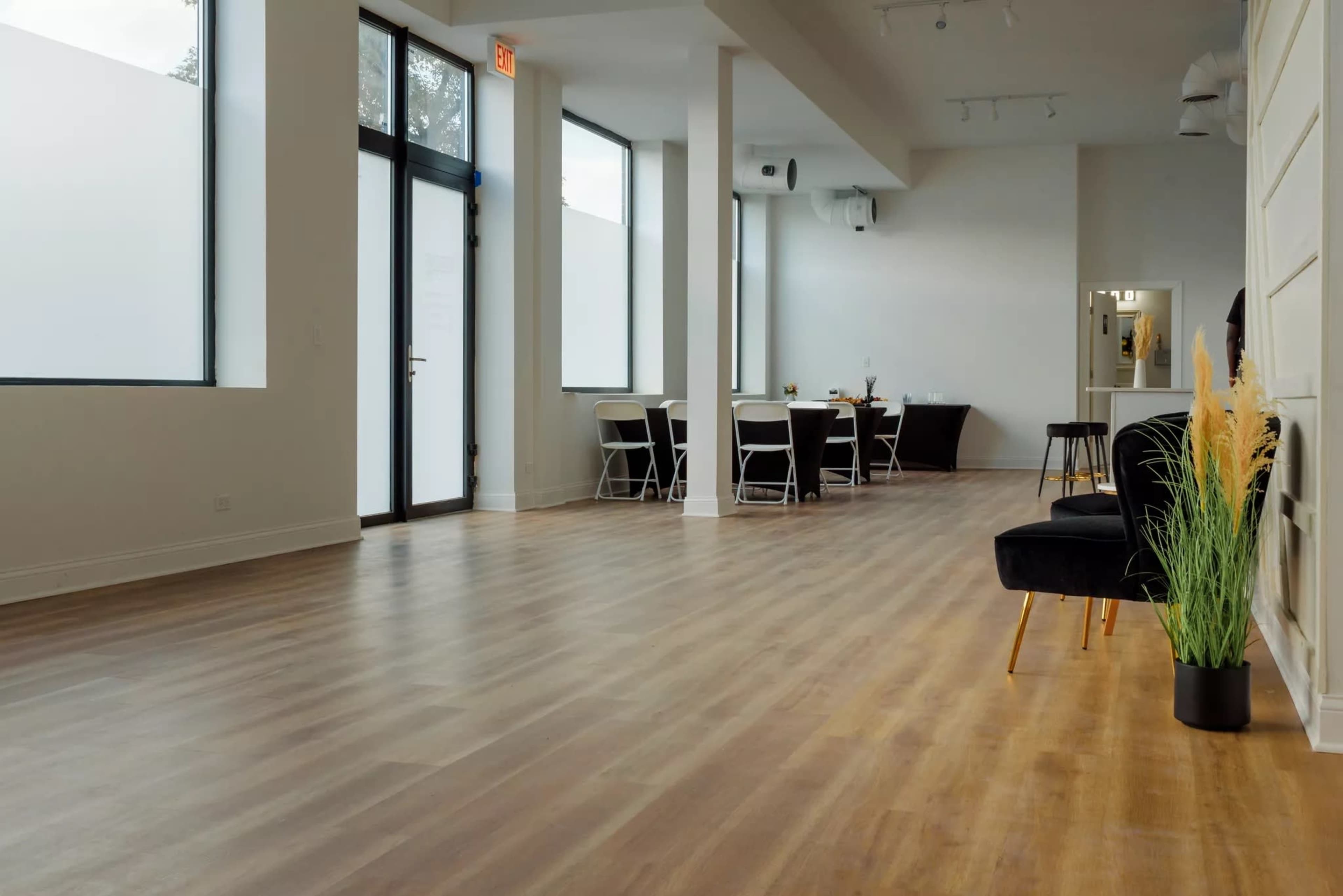 A spacious, airy interior with wooden flooring, large windows, and a seating area featuring chairs and a black chair against a wall.