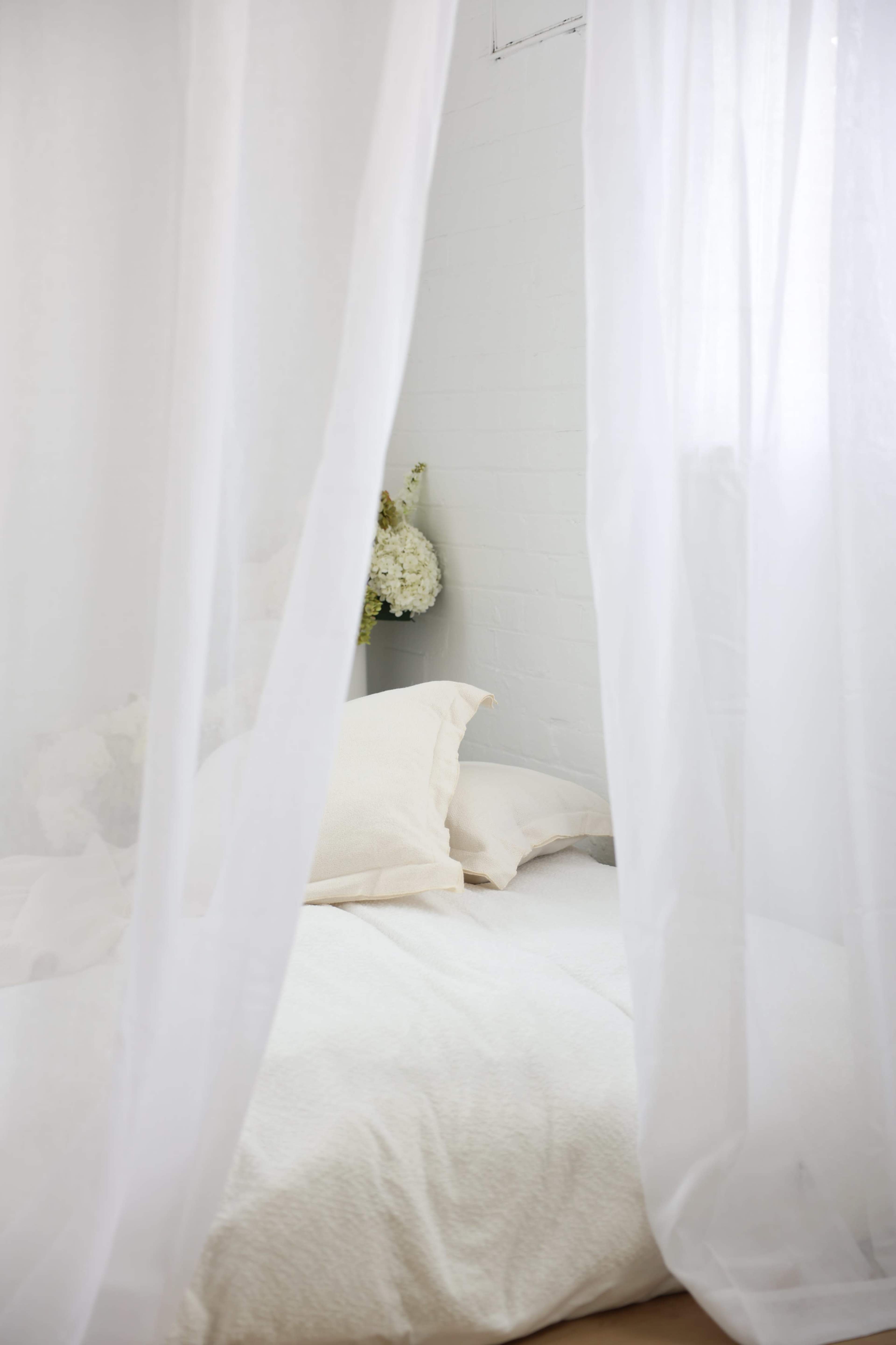 A bed with white bedding and decorative pillows is partially visible through sheer, white curtains.