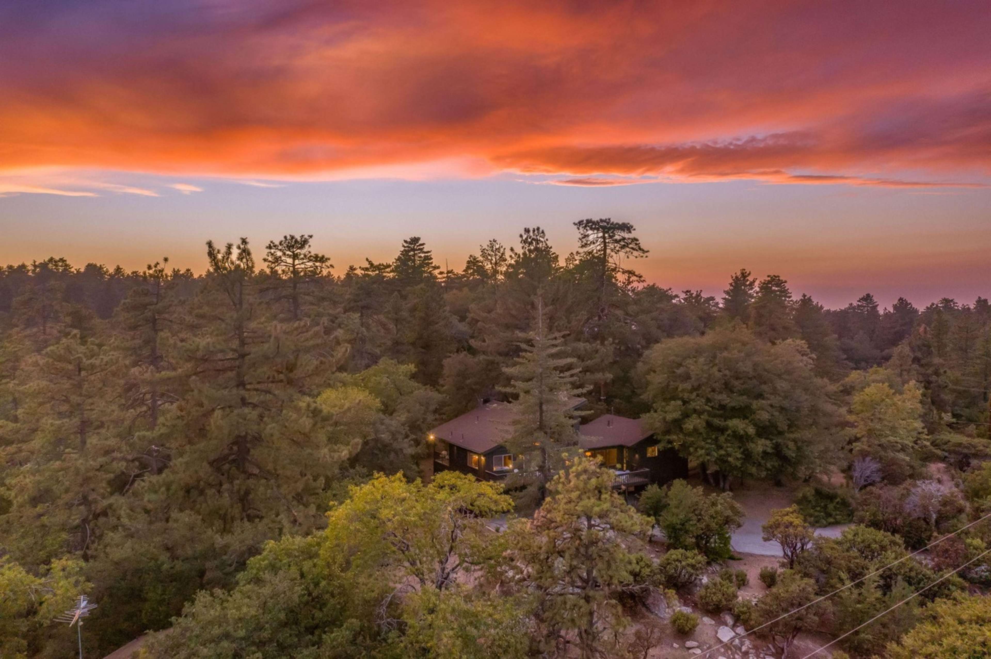 A cabin is nestled among tall trees under a vibrant sunset sky.