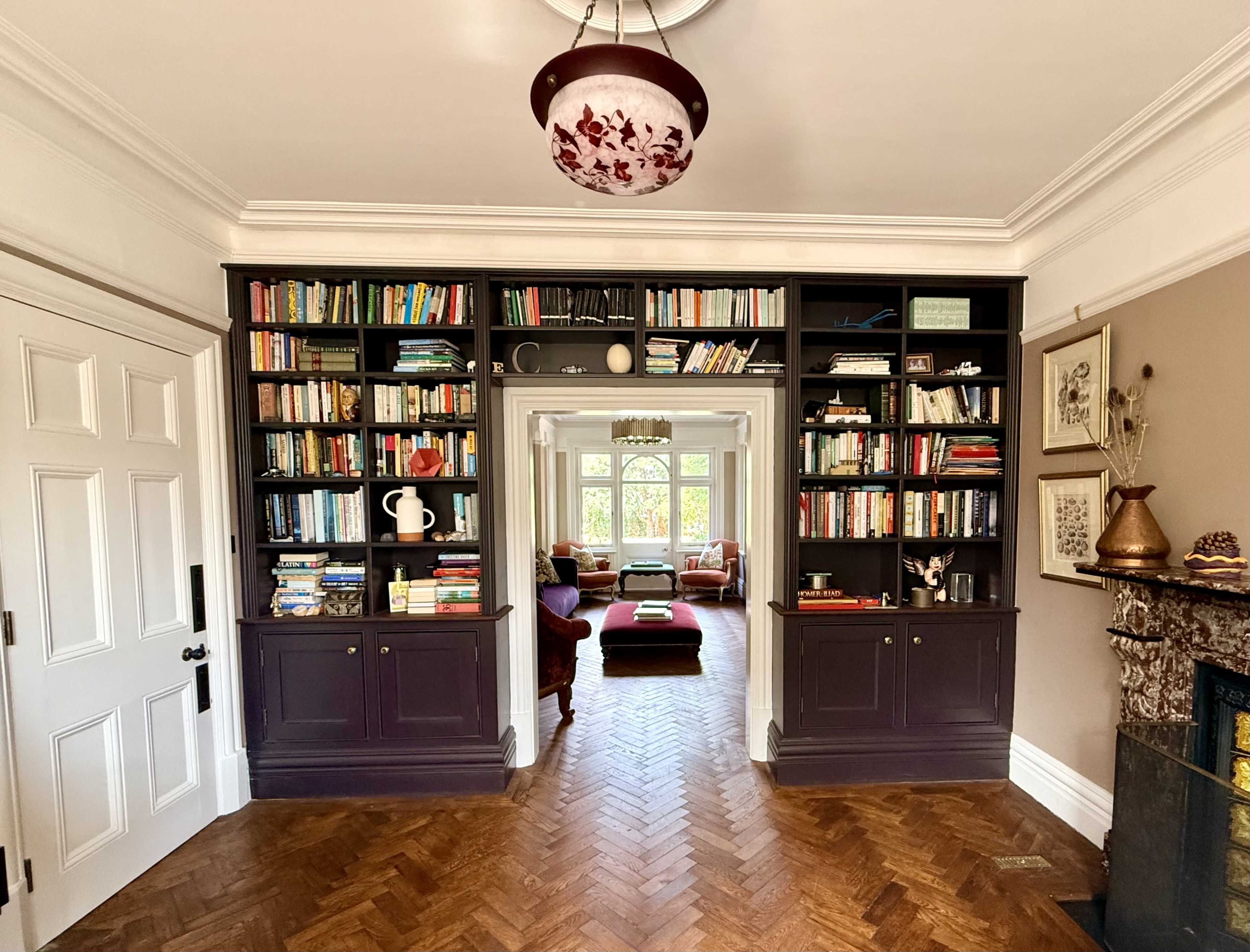 The image shows a cozy room with dark wooden bookshelves filled with books, a wooden floor, and natural light coming from large windows in the background.