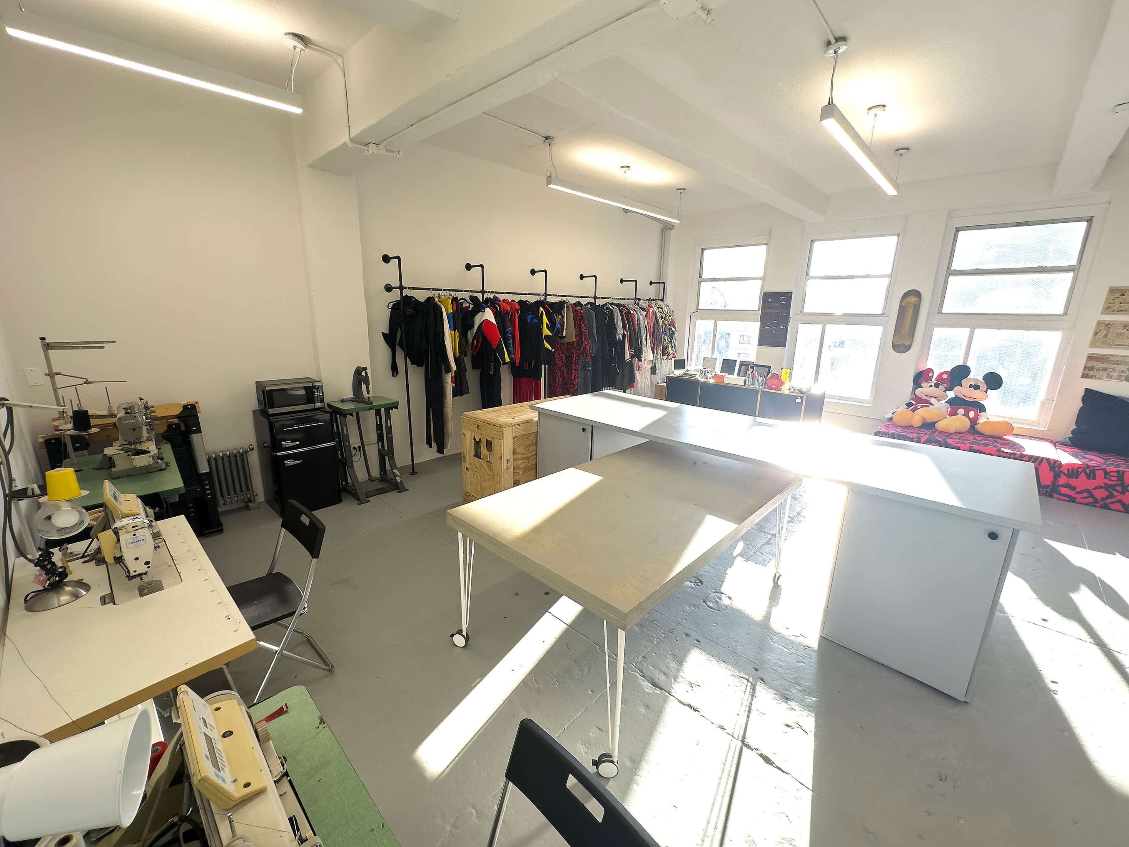 A bright sewing studio with work tables, a sewing machine area, and clothing racks filled with garments.
