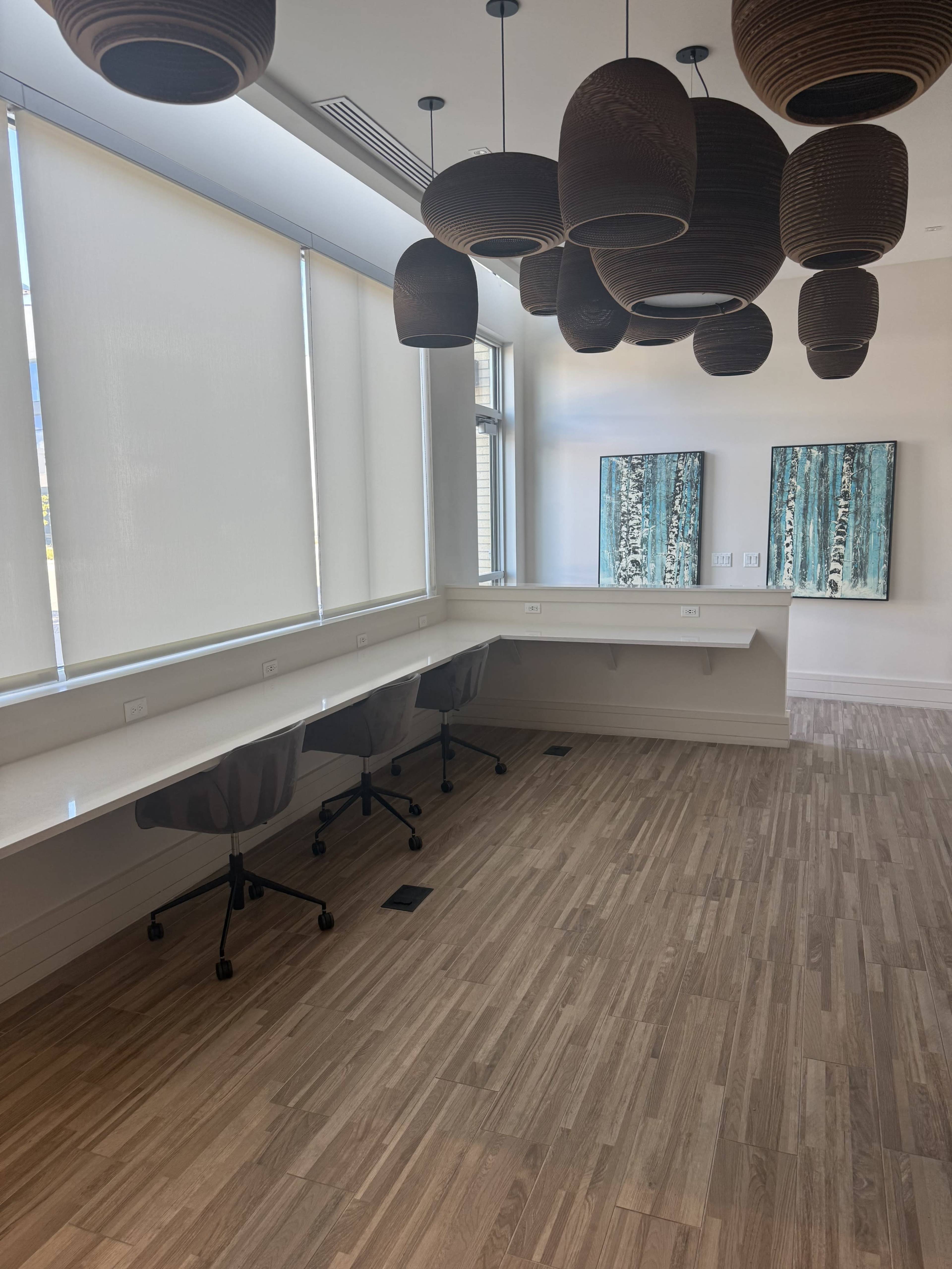 The image shows a modern office space featuring a light wooden floor, two ergonomic chairs at a white counter, and pendant lighting above, with two abstract artworks on the wall.