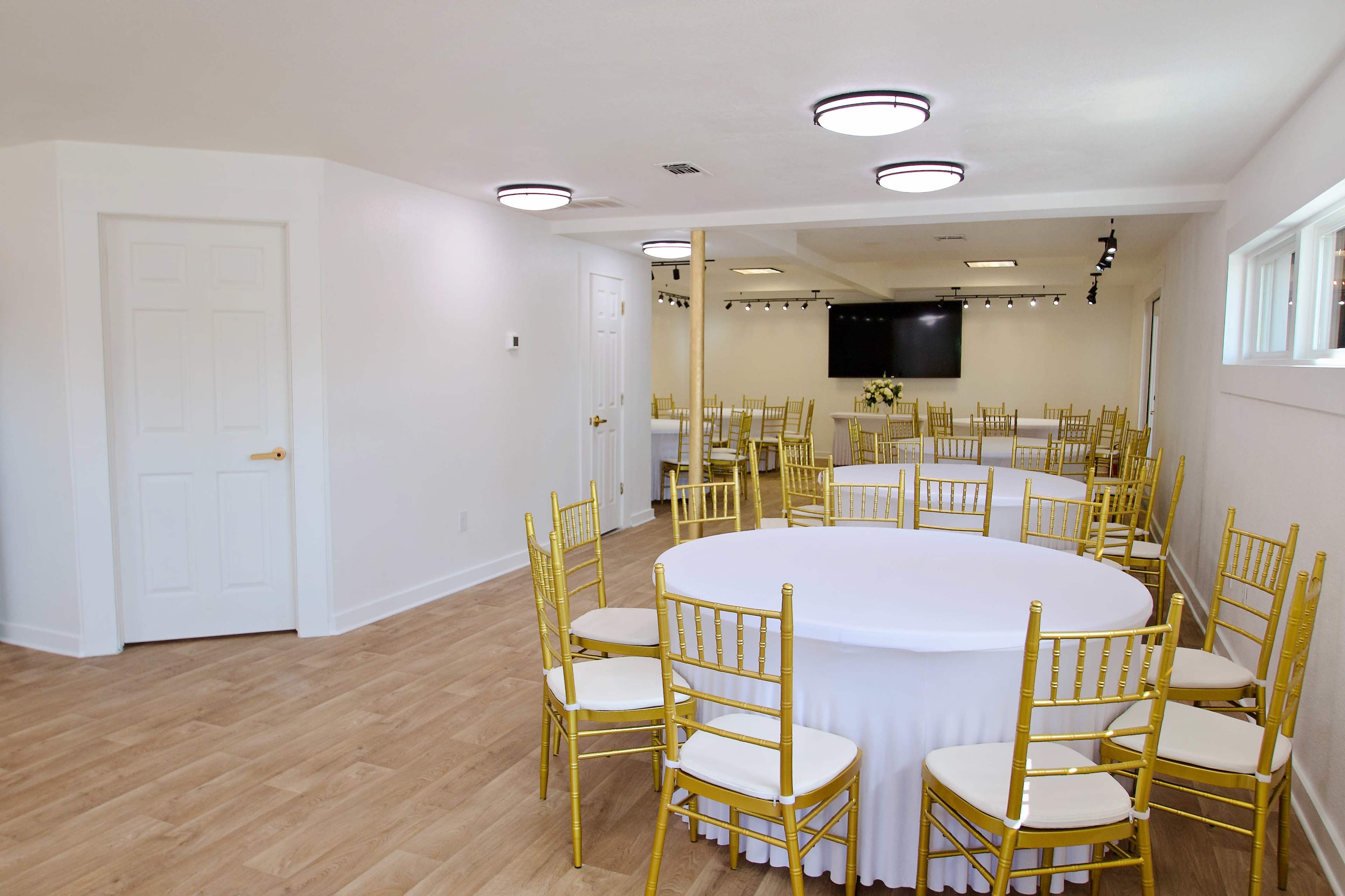 The image shows a spacious event room with multiple round tables set with white tablecloths and gold chairs, illuminated by overhead lights.