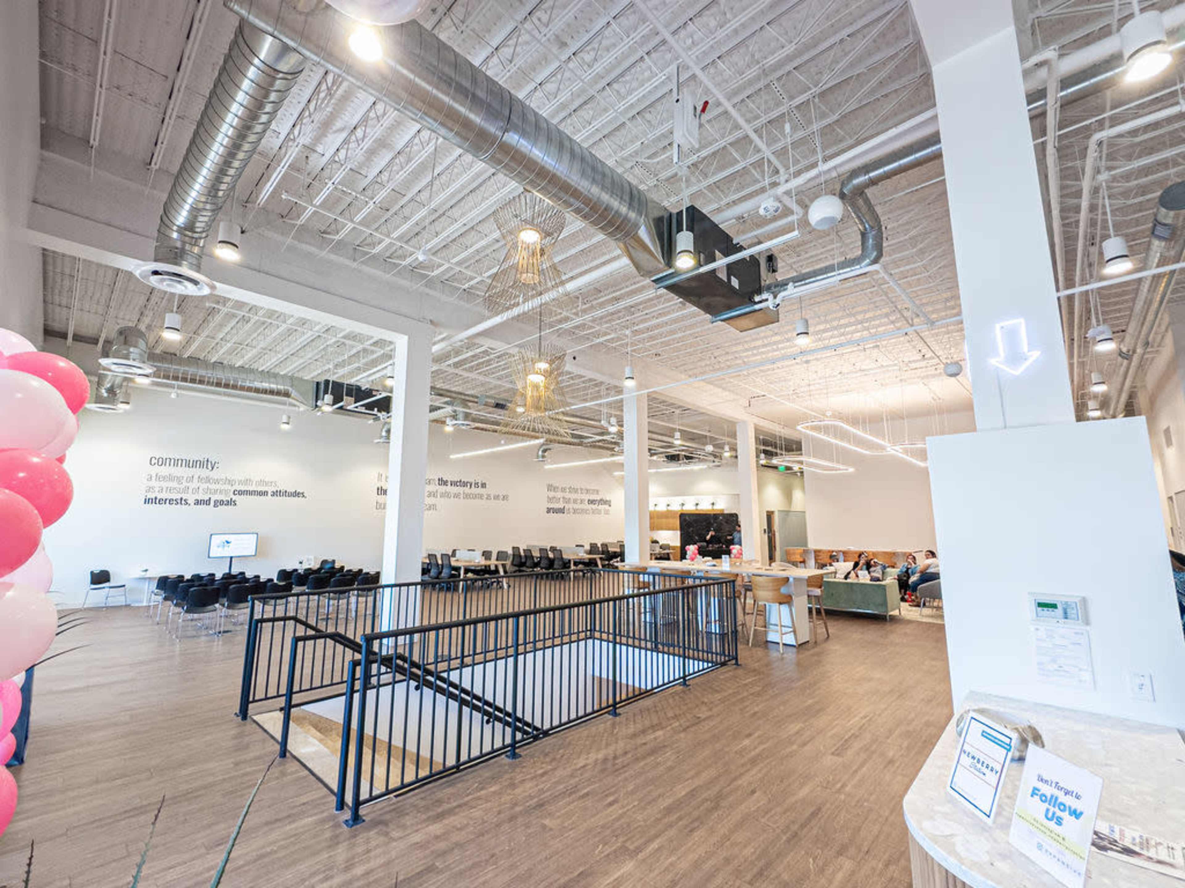 The image shows a modern indoor space with high ceilings, featuring a mix of seating areas, a staircase, and walls adorned with large text, all under bright lighting.