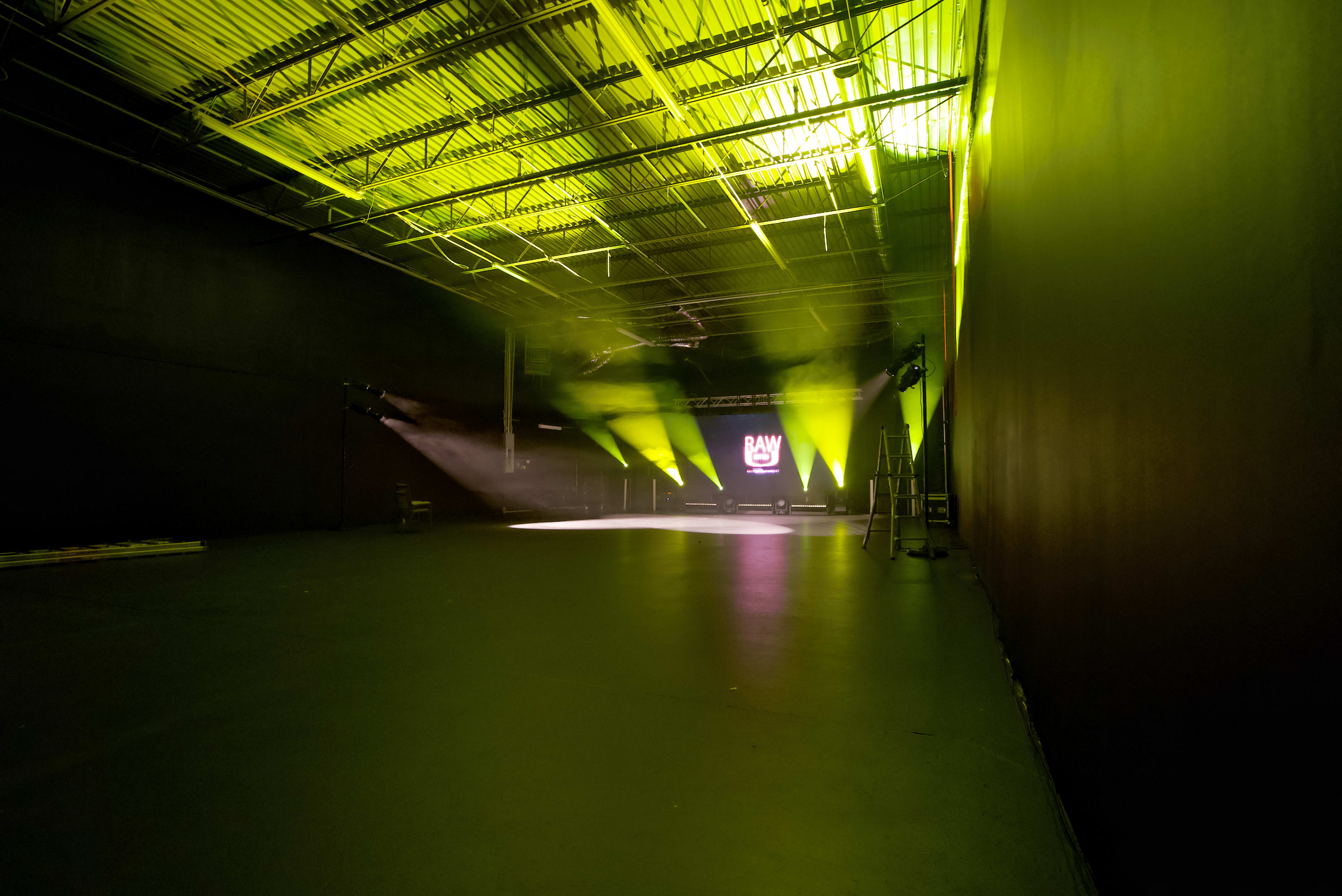 A spacious, dimly lit warehouse features green and yellow stage lights illuminating a blank performance area, with a logo displayed on a screen in the background.