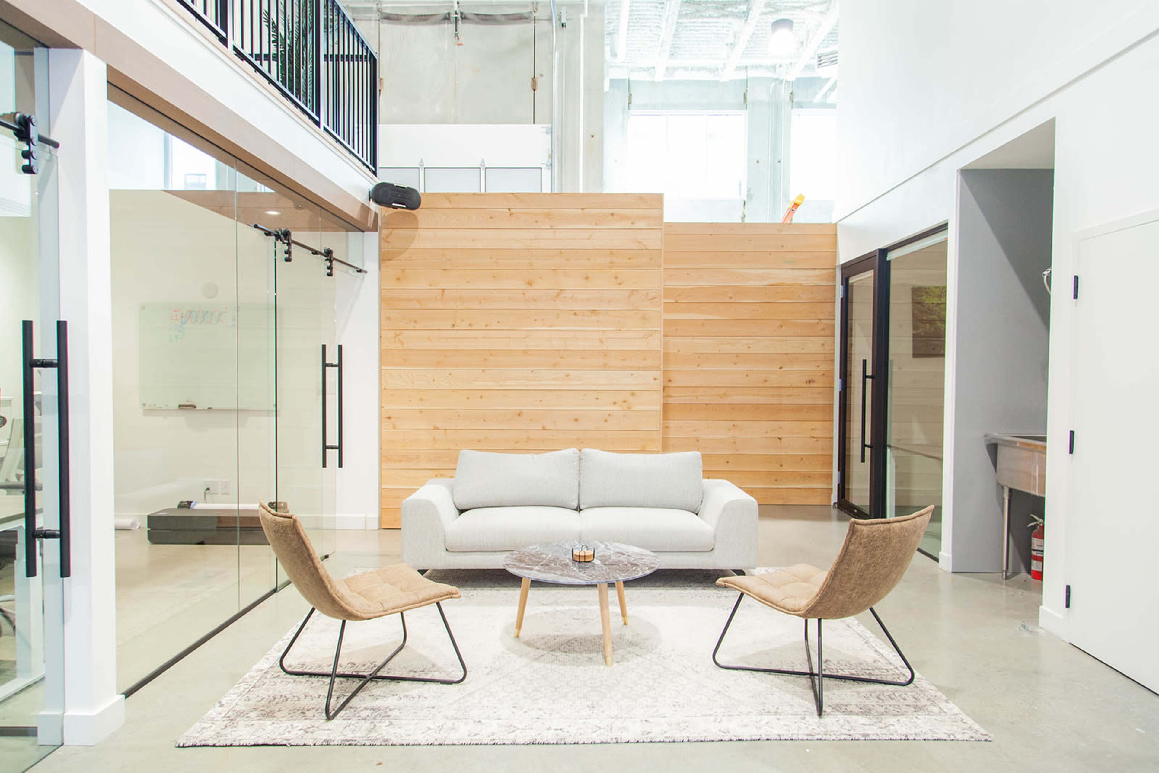 A modern seating area featuring a light gray sofa and two brown armchairs arranged around a circular coffee table on a patterned rug within a bright, minimalist space.