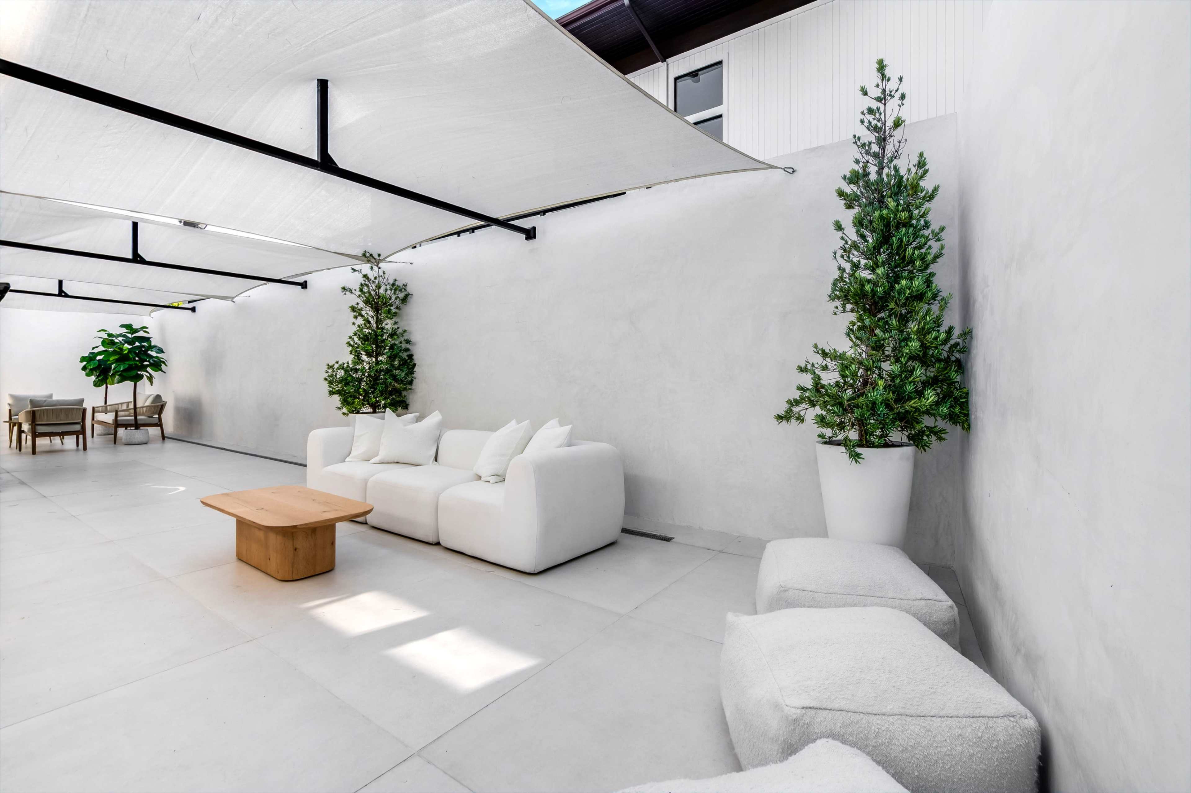 The image shows a minimalist indoor space featuring white walls, a light-colored sofa, a wooden coffee table, and potted greenery.