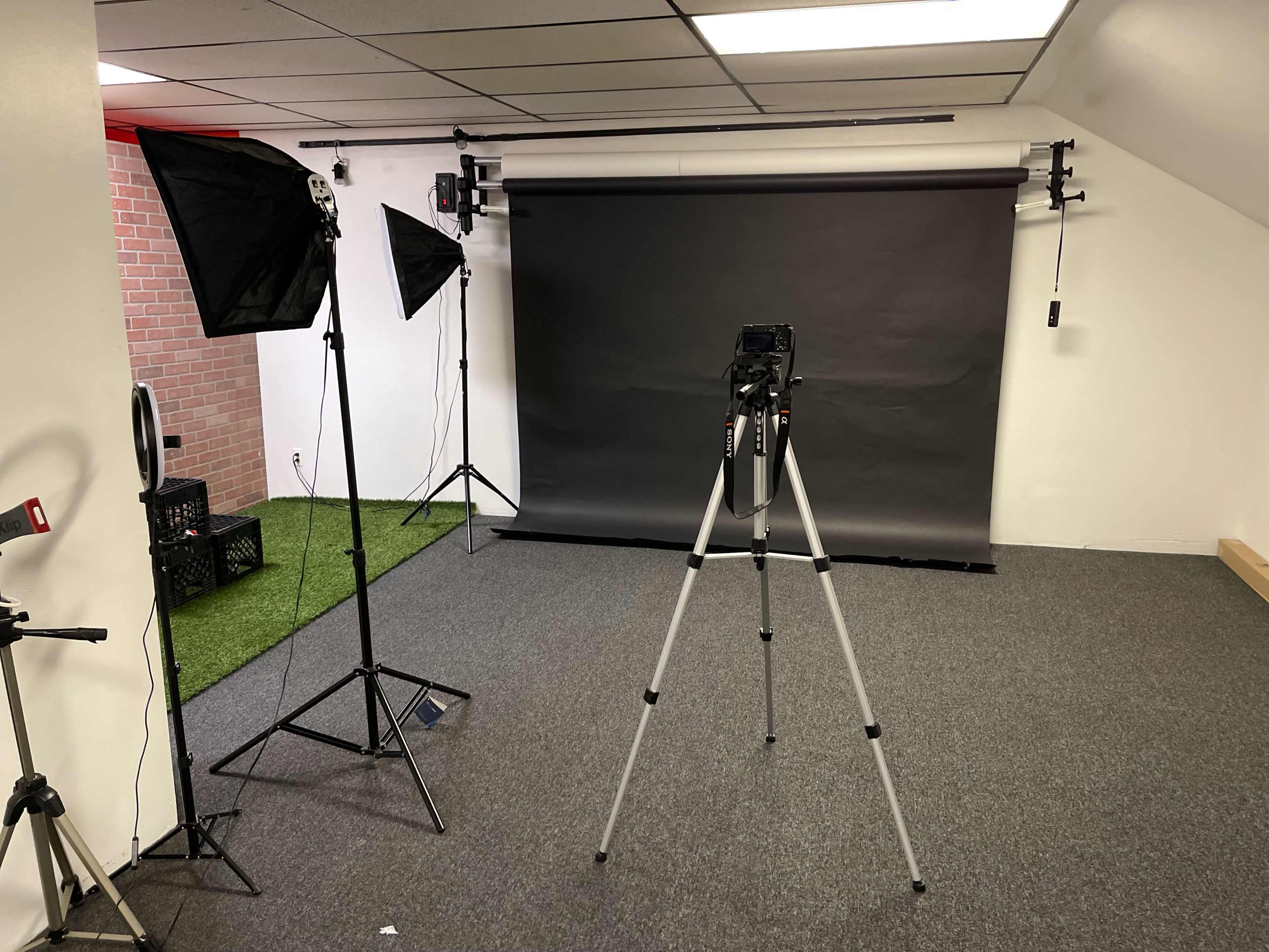 A photography studio setup with a black backdrop, a camera on a tripod, and two softbox lights on either side.