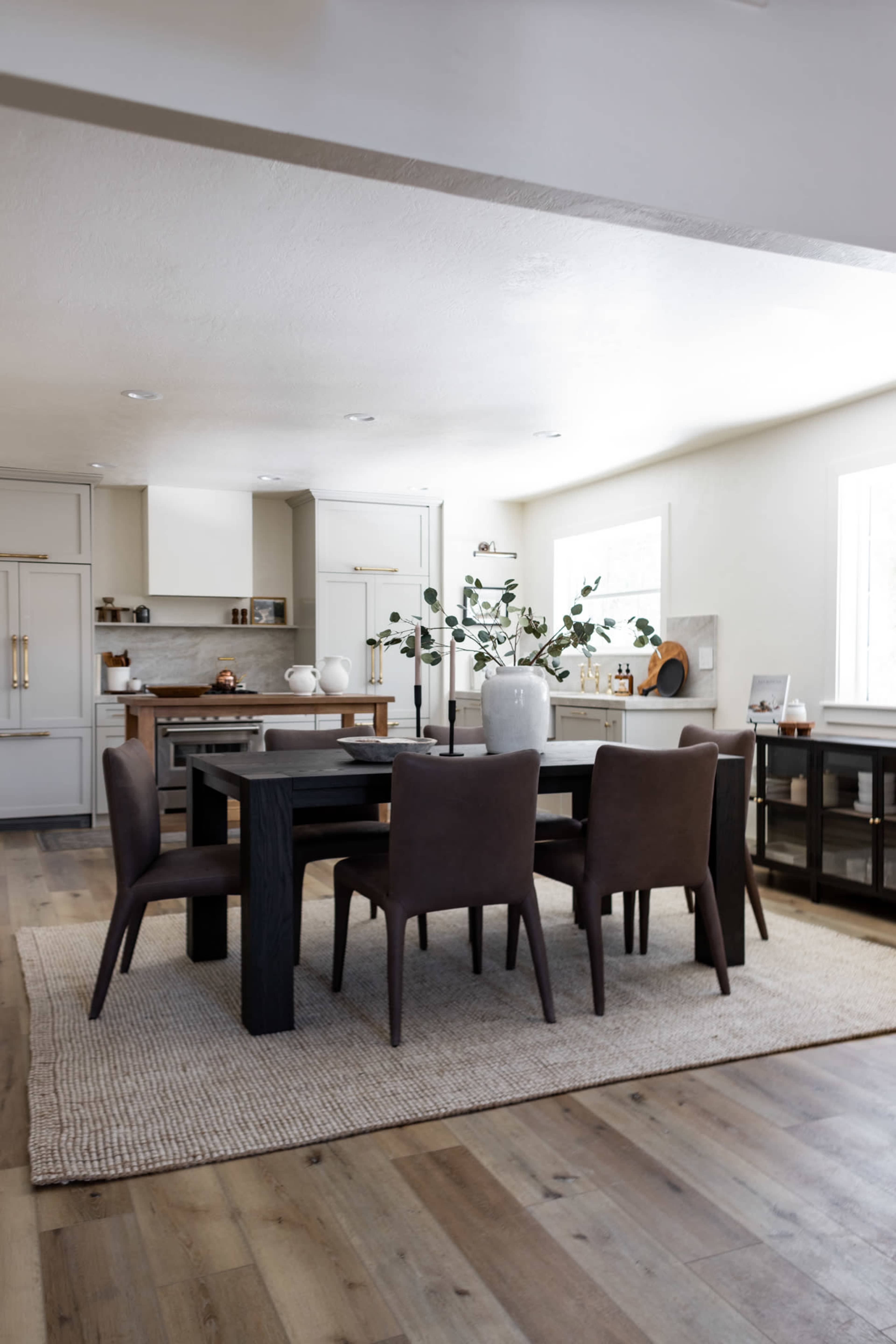 A modern dining room features a dark wooden table surrounded by four brown chairs, with a large vase and greenery on the table, set on a woven area rug.