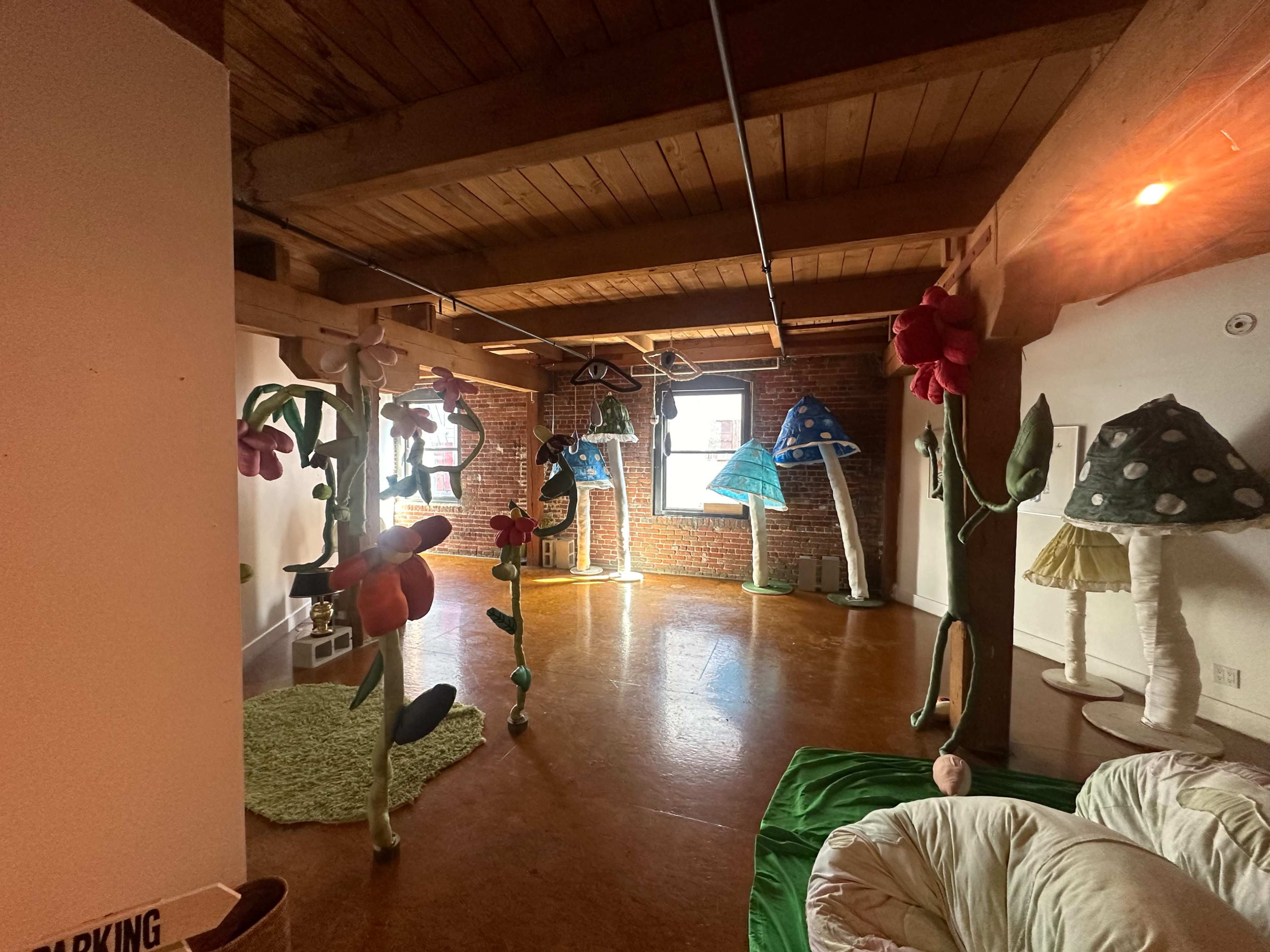 The room features large, colorful fabric sculptures resembling flowers and mushrooms, set against a backdrop of exposed brick walls and wooden beams.