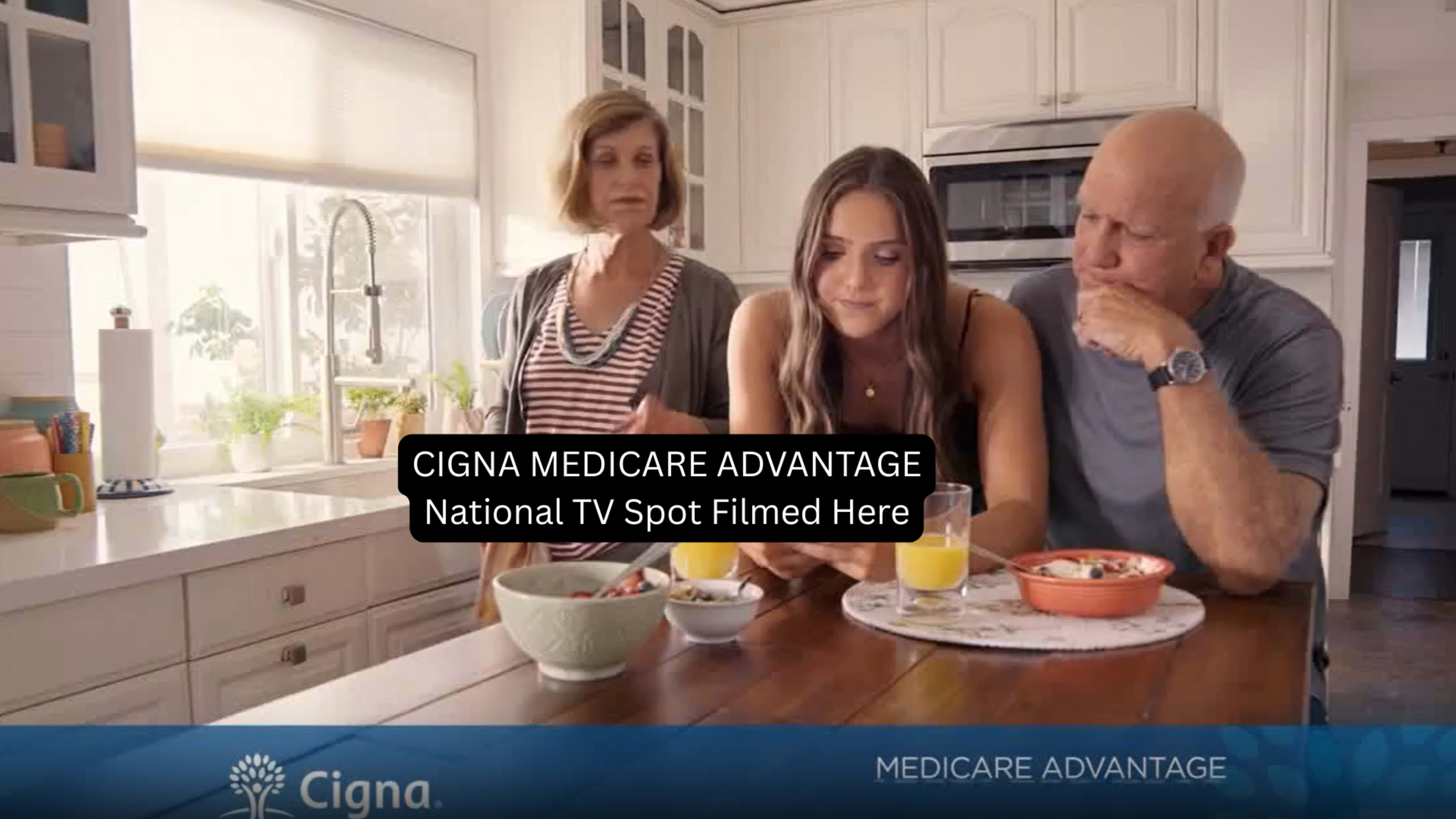 A woman sits at a kitchen table looking at her phone, while two adults watch her with interest, and breakfast items are spread out before them.