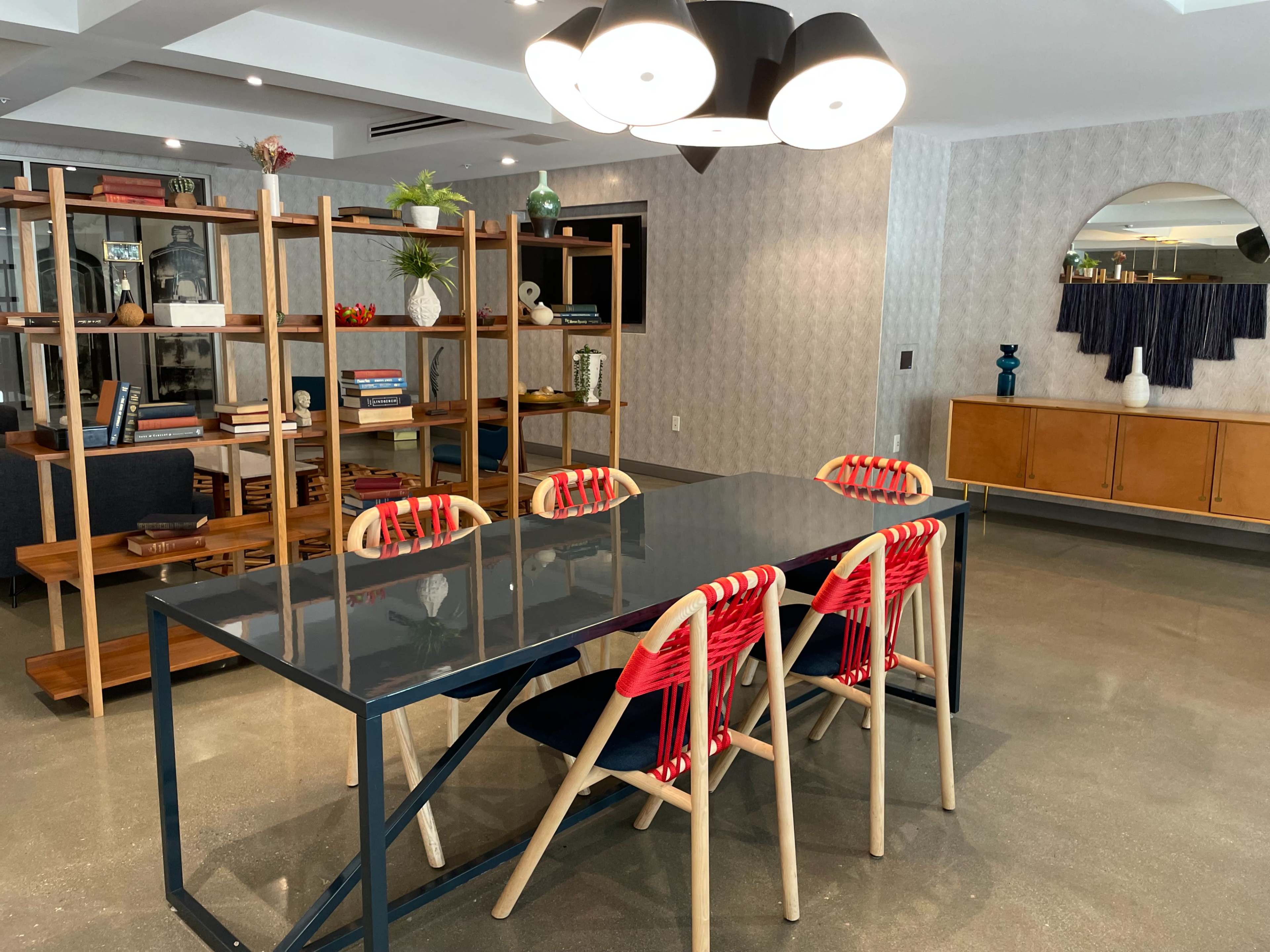 A modern dining area featuring a rectangular blue table surrounded by chairs with red woven backrests, and a wooden shelving unit in the background.