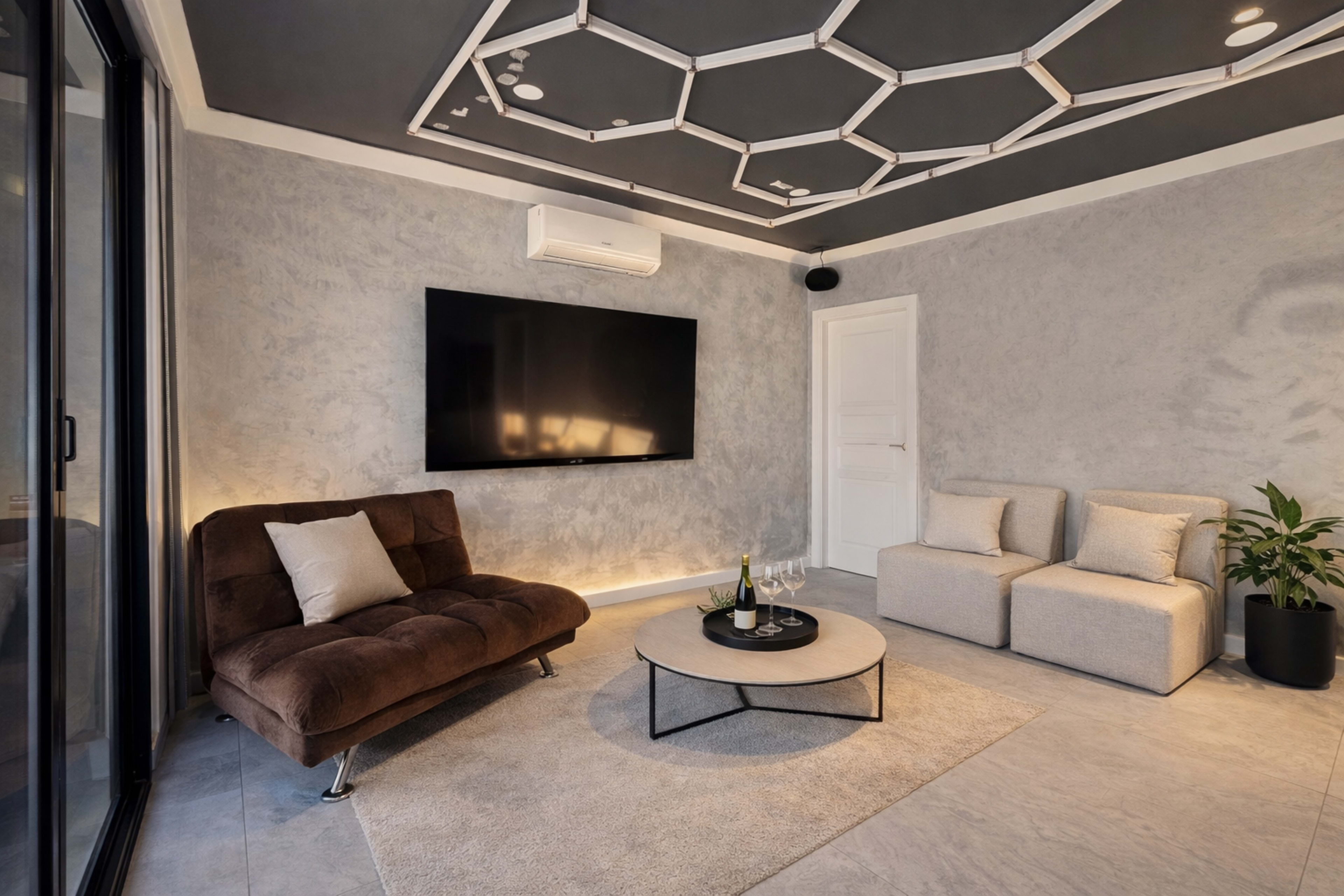 The image shows a modern living room with a brown sofa, a coffee table, and a gray rug against a textured wall, featuring a large wall-mounted television and a plant in a corner.