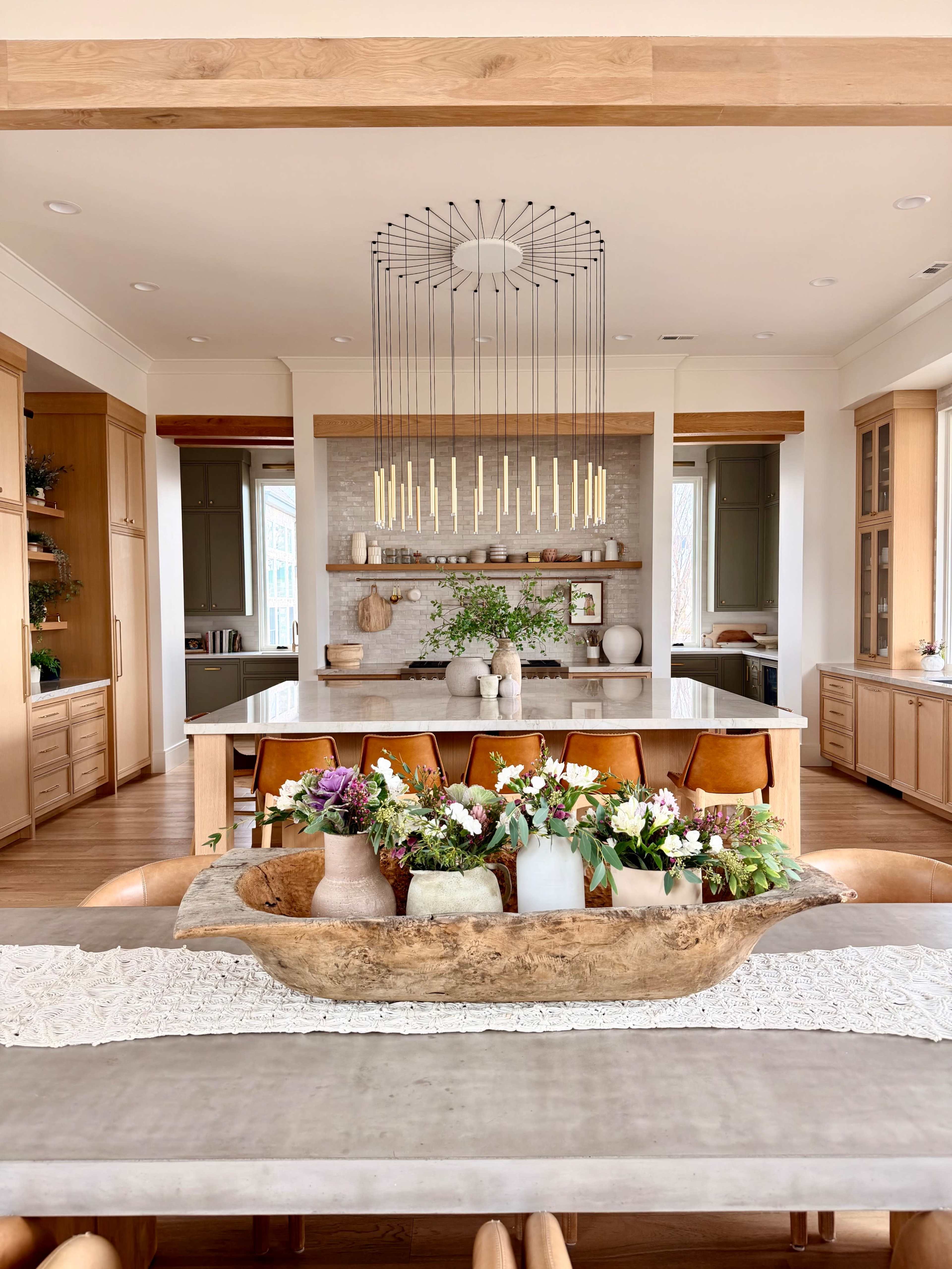 A modern kitchen features a large island with bar stools, pendant lighting, and decorative vases filled with flowers on a dining table.