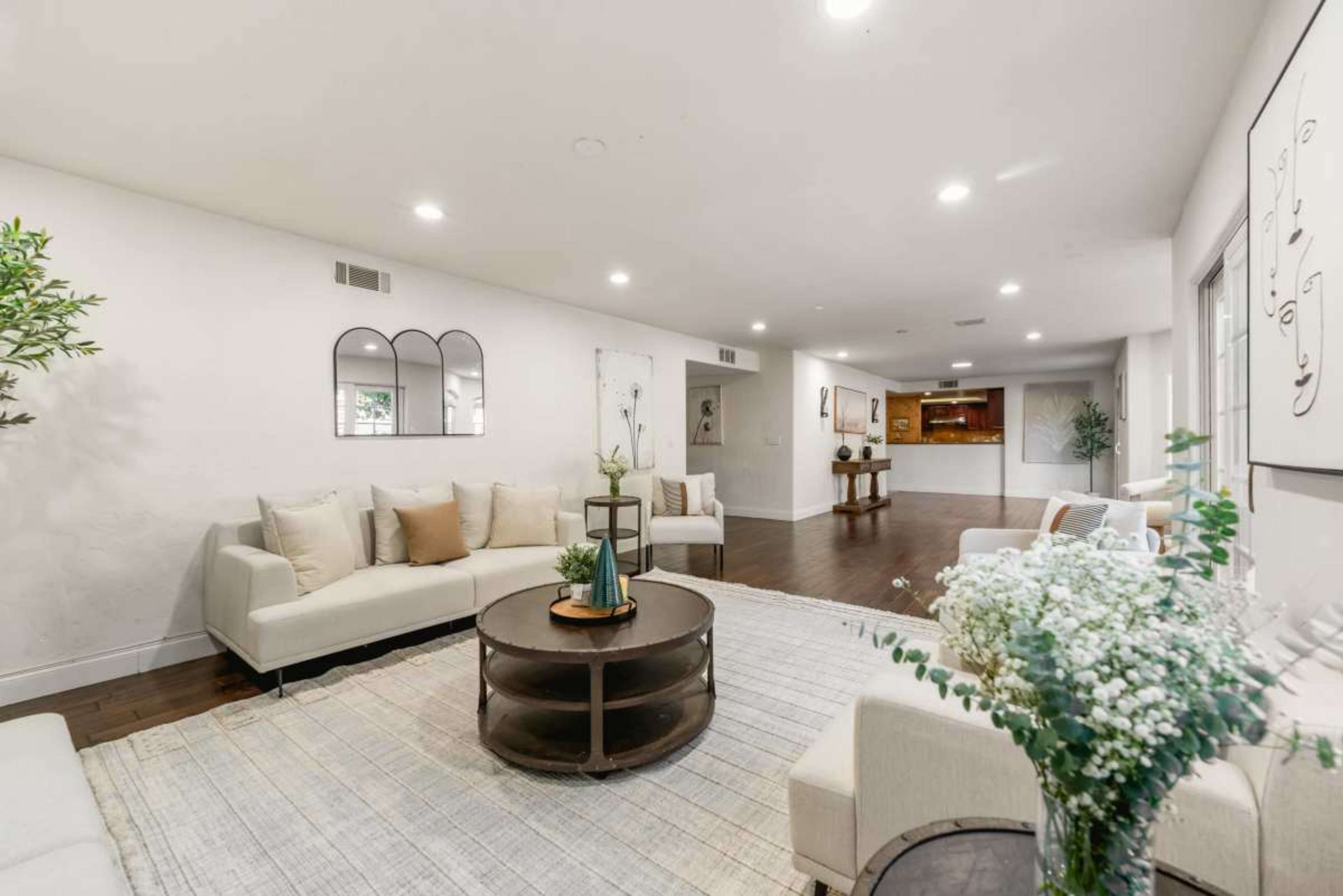 The image shows a spacious living room with sectional sofas, a round coffee table, and decorative elements like plants and artwork on the walls.