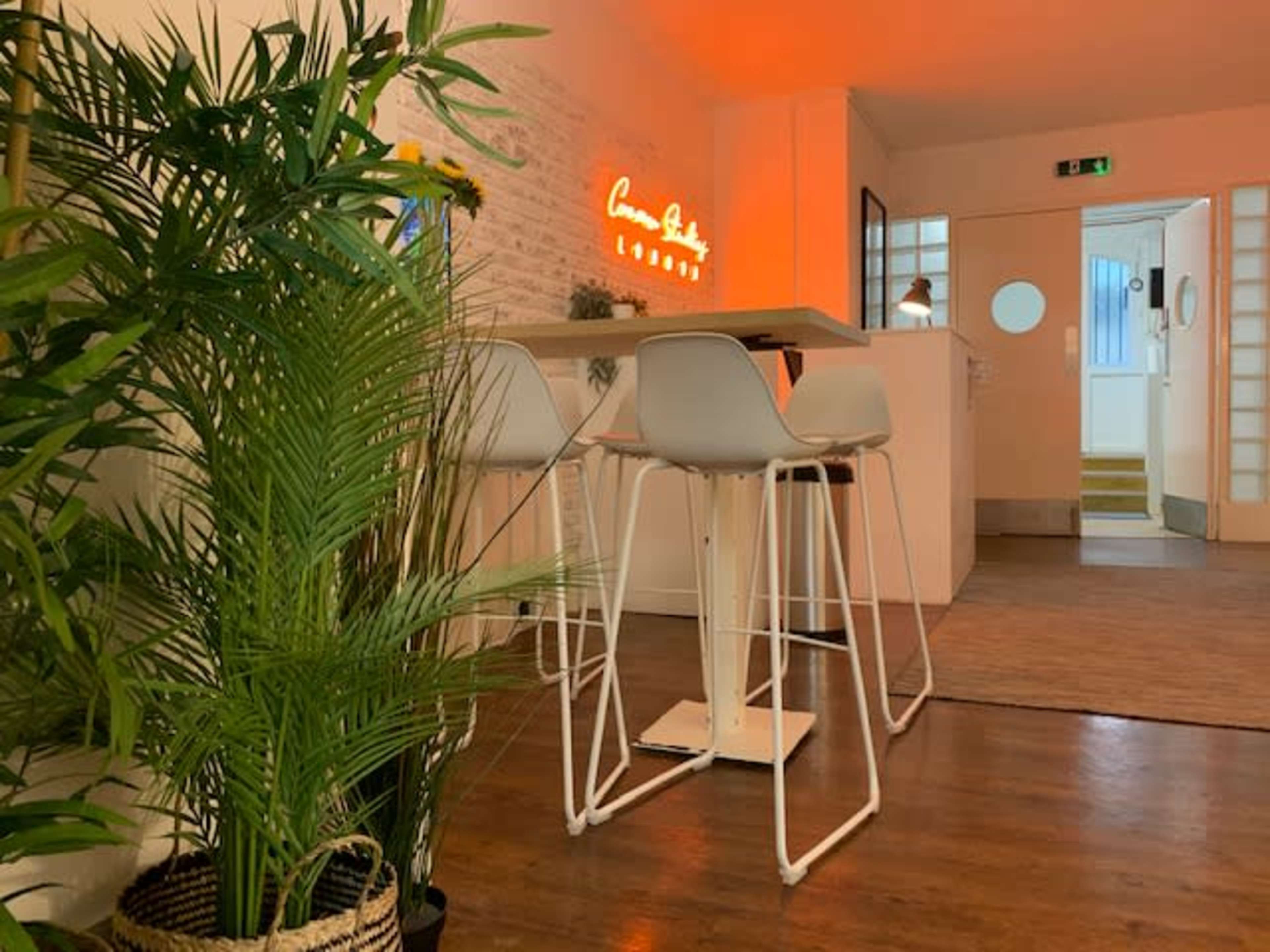A modern lounge area with a high table and stools beside a potted plant, illuminated by a glowing neon sign.