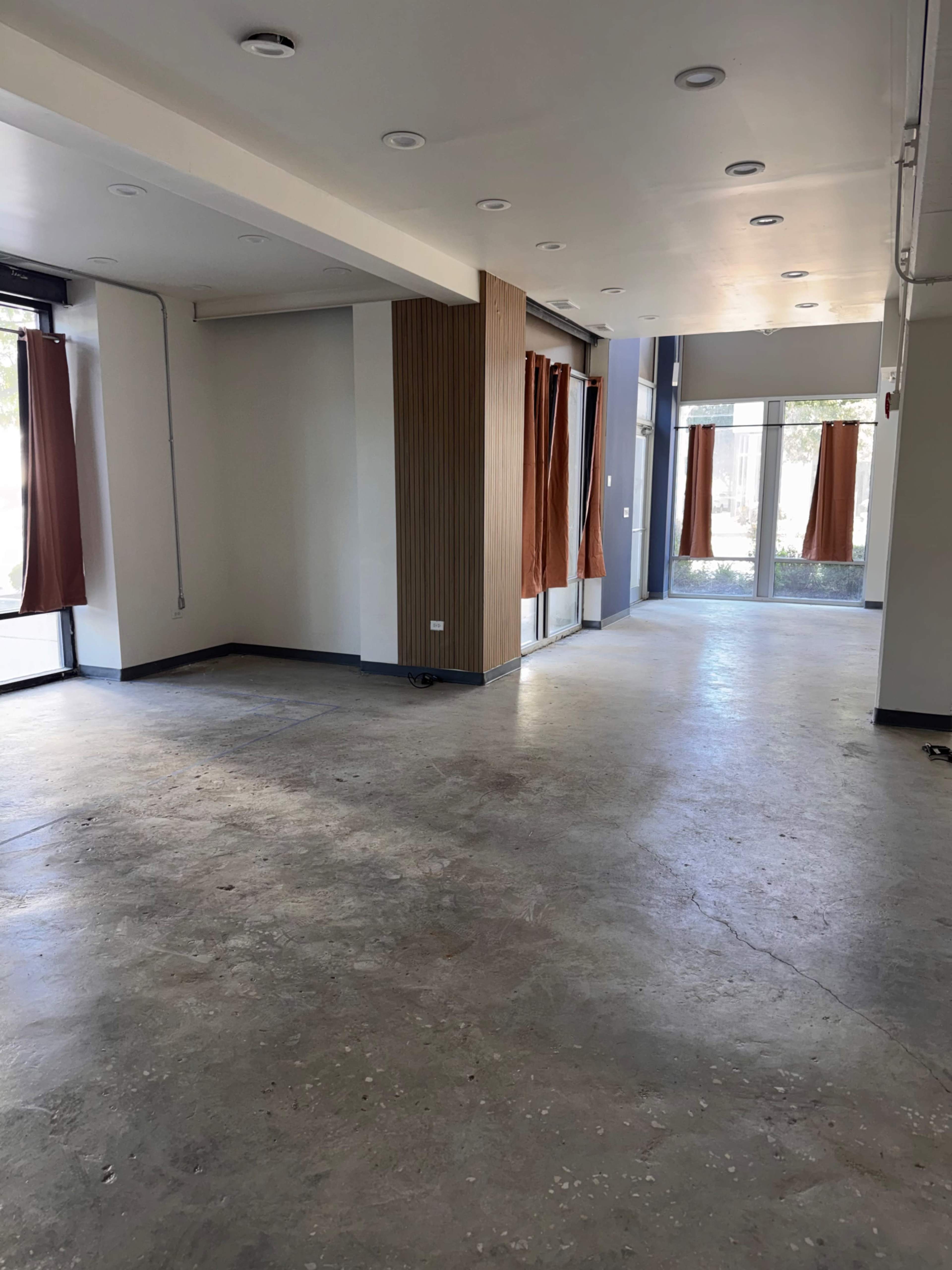The image shows an empty, well-lit interior space with large windows and brown curtains, featuring concrete flooring and minimal furnishings.