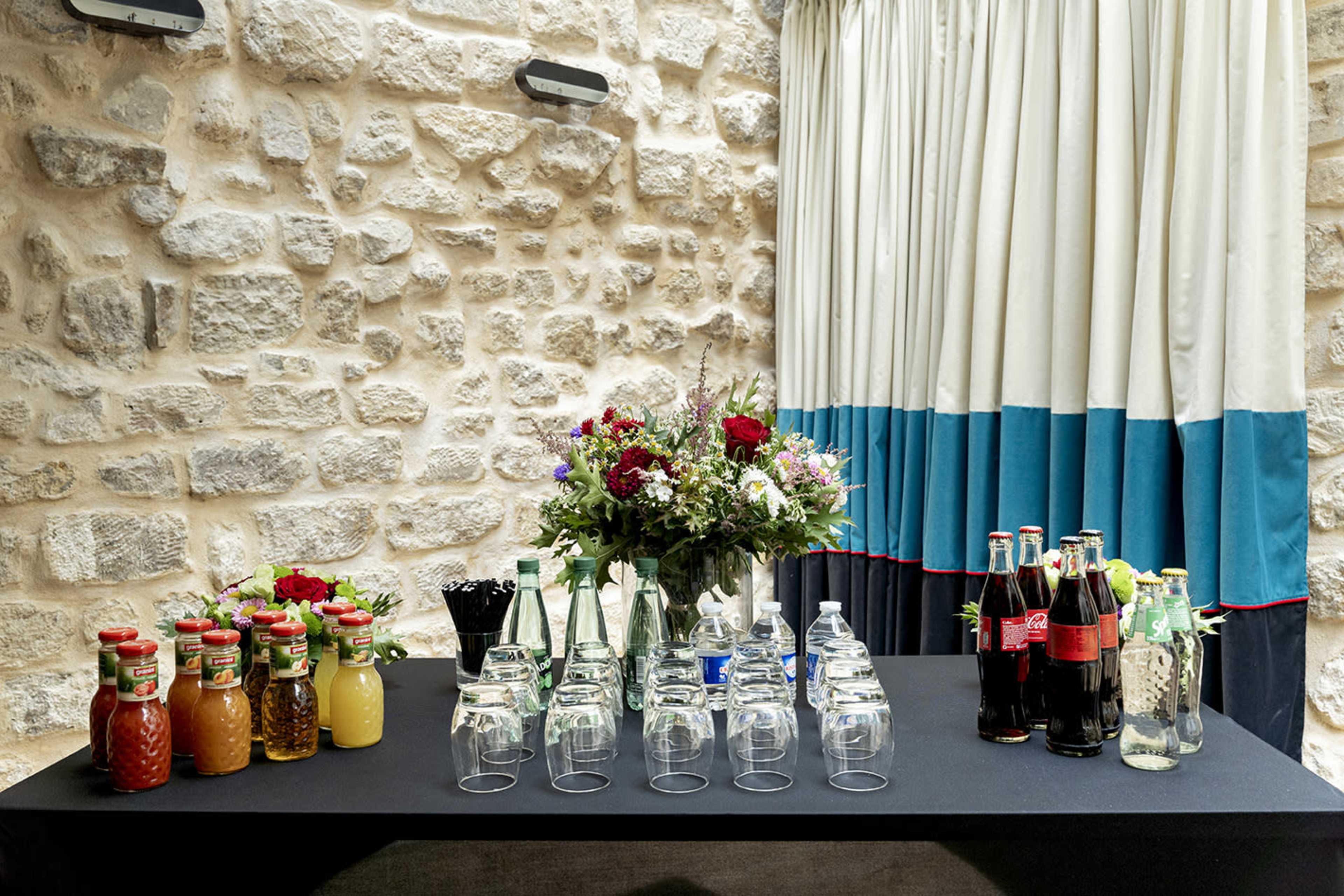 A table is set against a stone wall, displaying various bottles of juices, sodas, and glasses, alongside a floral arrangement.