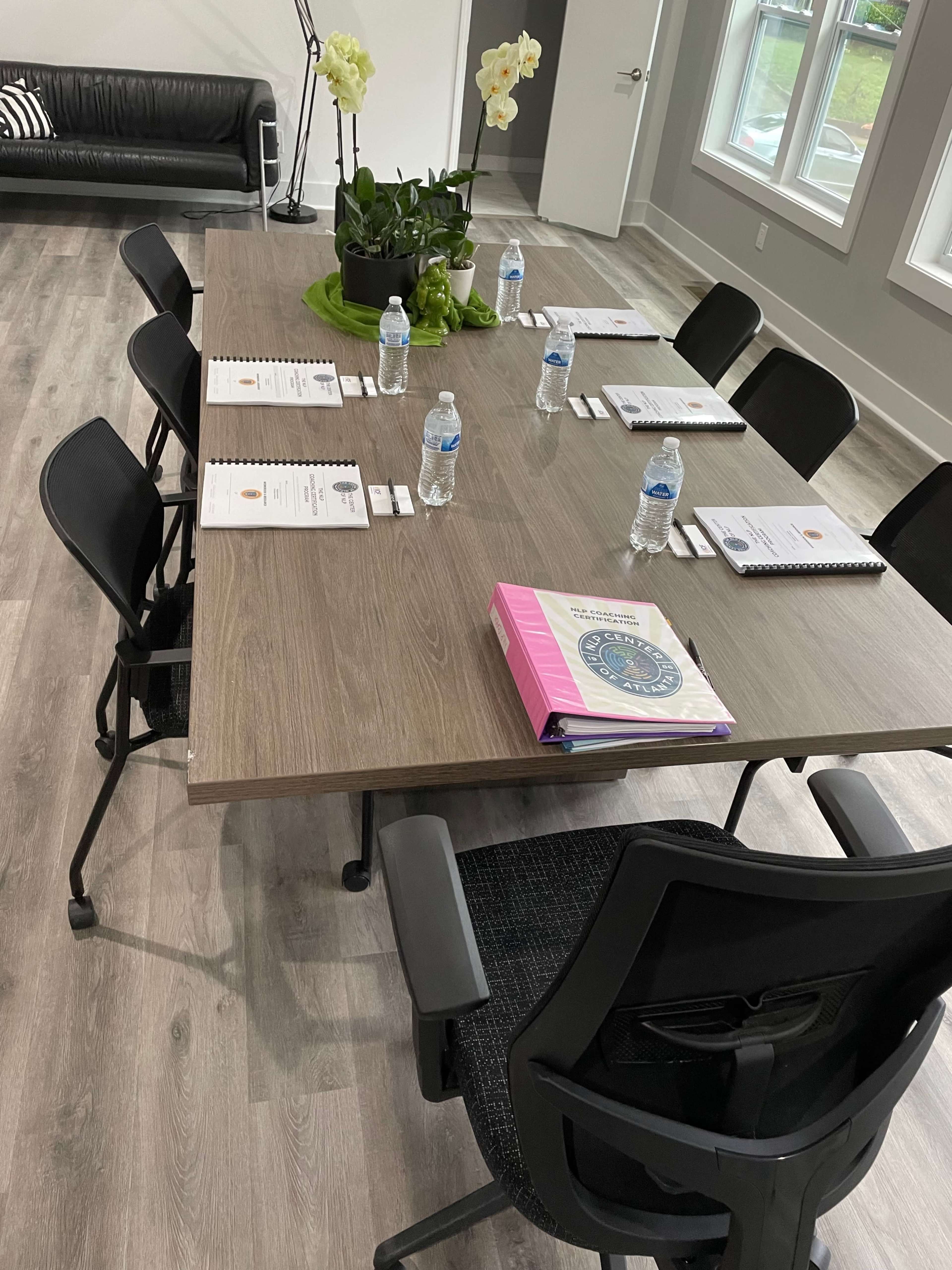 A modern meeting room features a large rectangular table with chairs, water bottles, notebooks, and a potted plant, set against a backdrop of large windows and hardwood flooring.