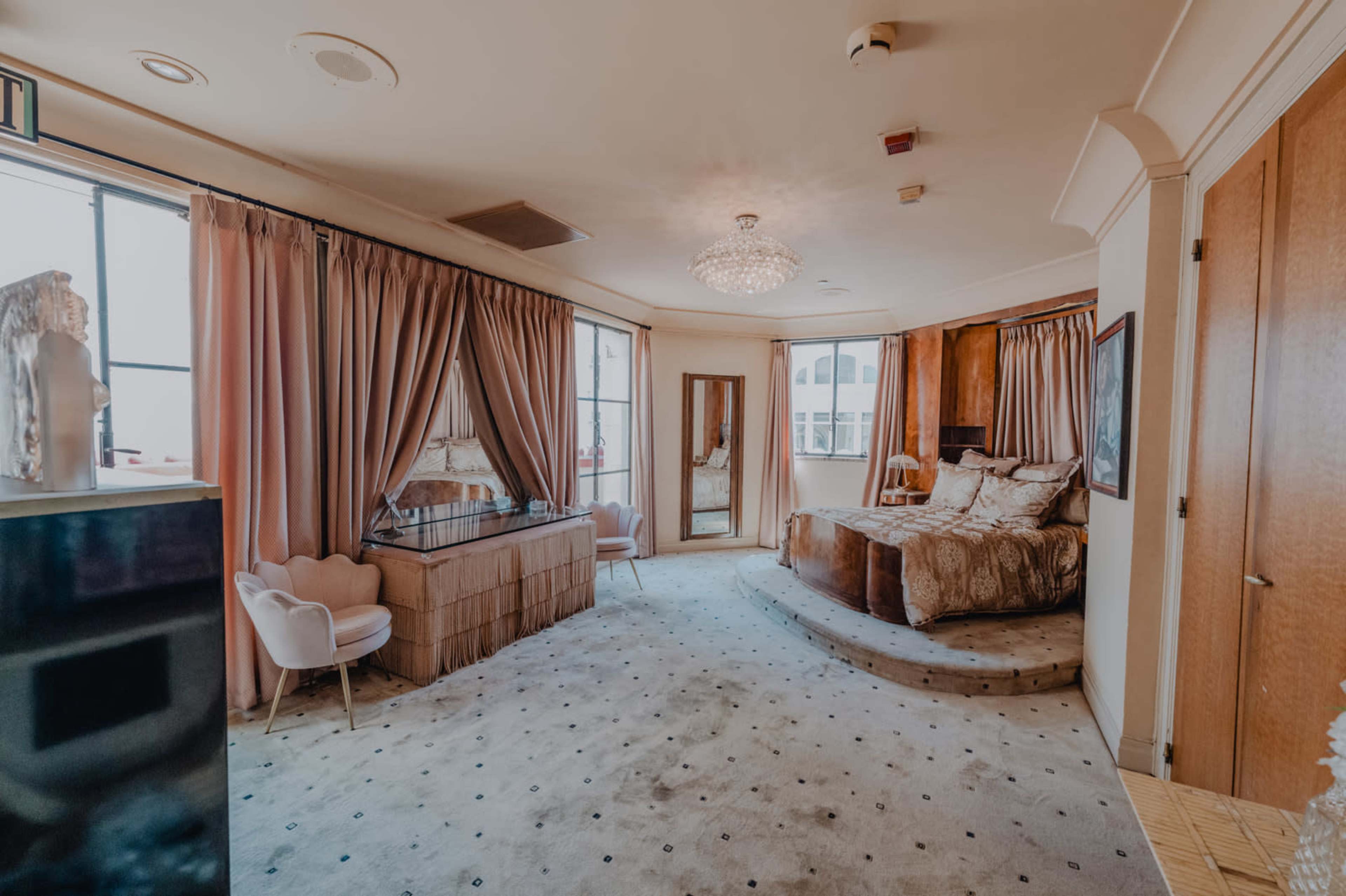 The image shows a luxurious bedroom with a large bed, elegantly draped curtains, a chandelier, and a vanity area with seating.