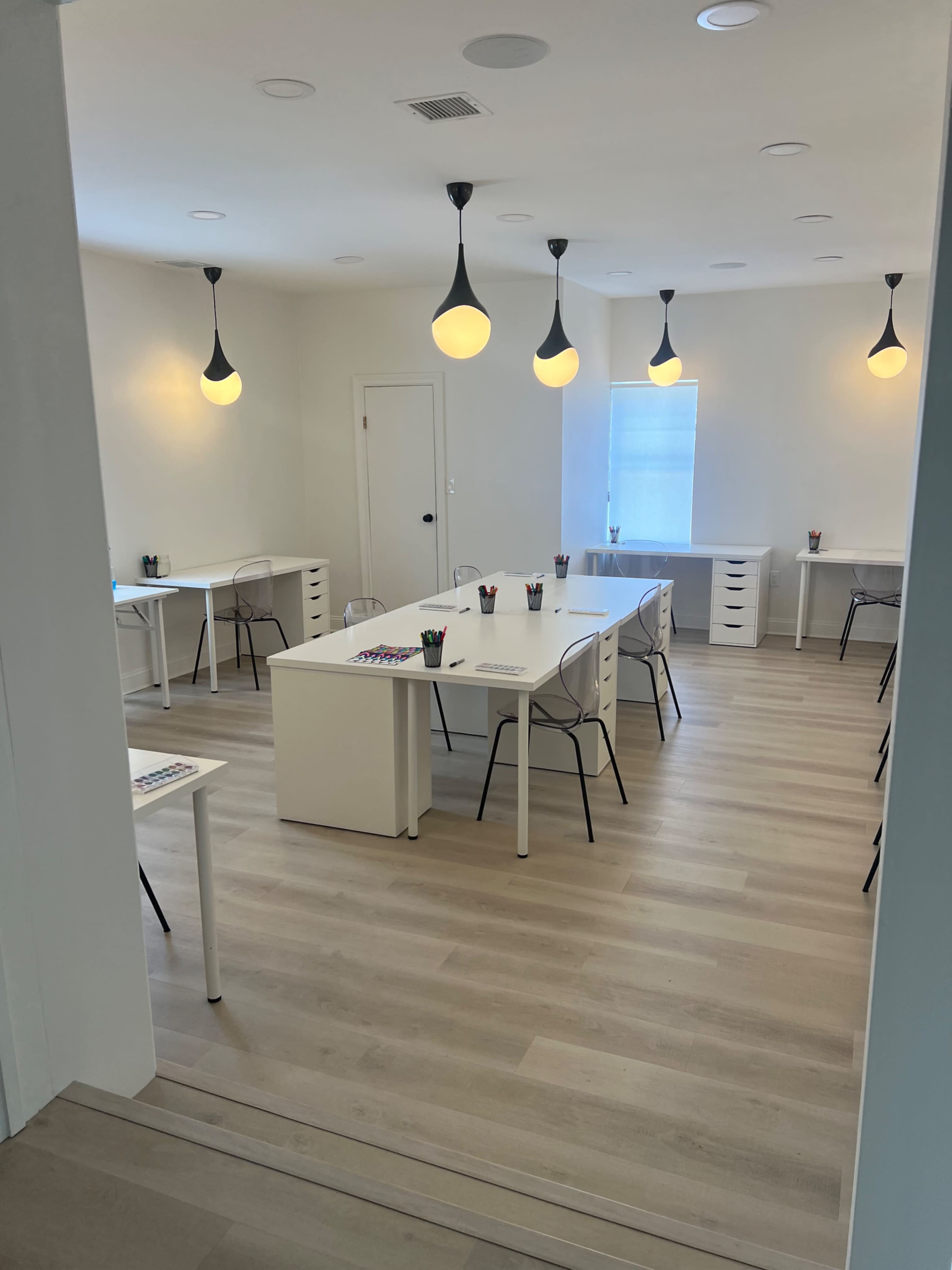 A bright, minimalist room with white tables, chairs, and pendant lighting, arranged in a classroom or meeting style.