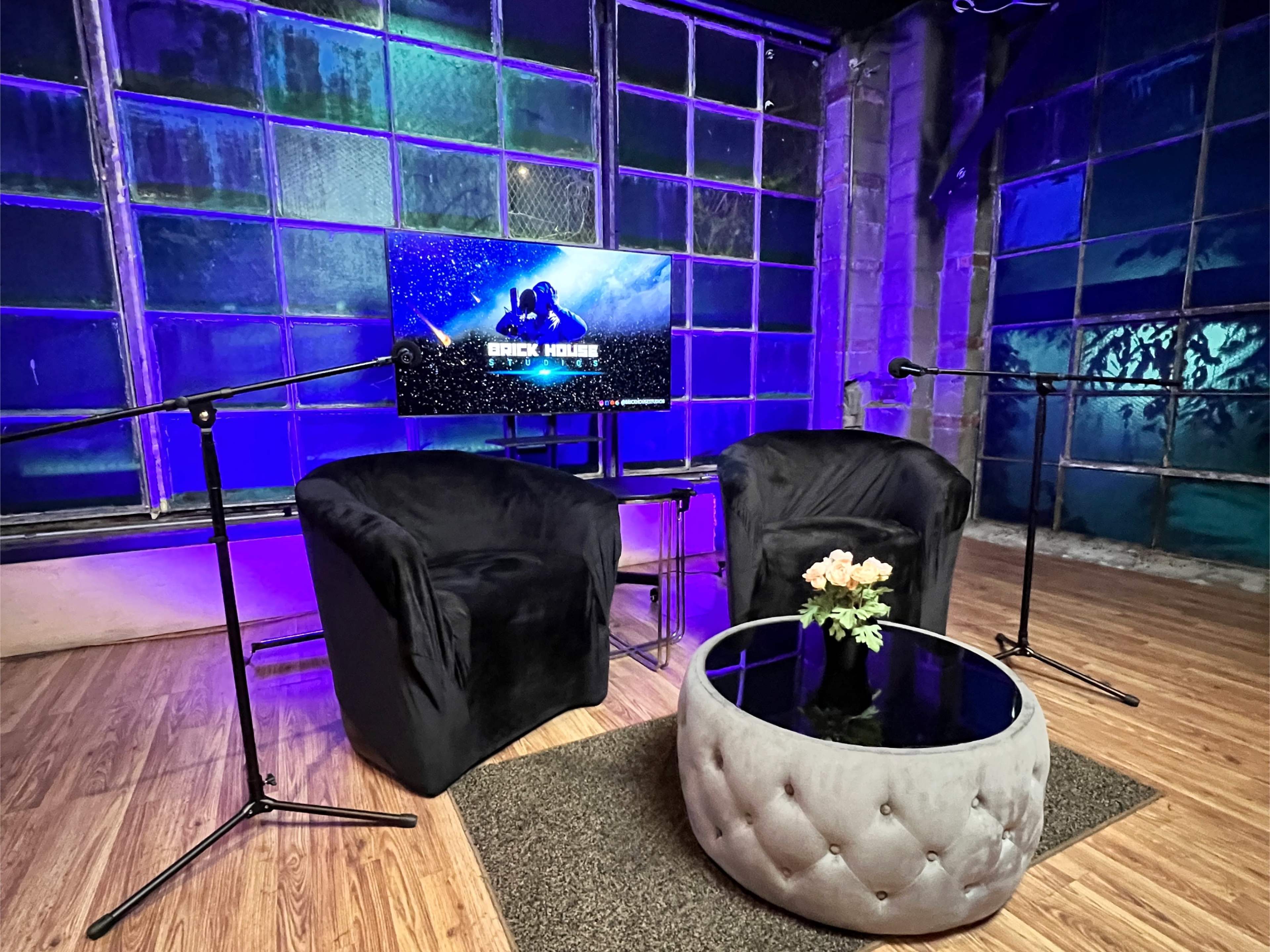 A podcast or interview setup featuring two black chairs, a glass coffee table with flowers, and a screen displaying a graphic, all set against a backdrop of large, illuminated windows.
