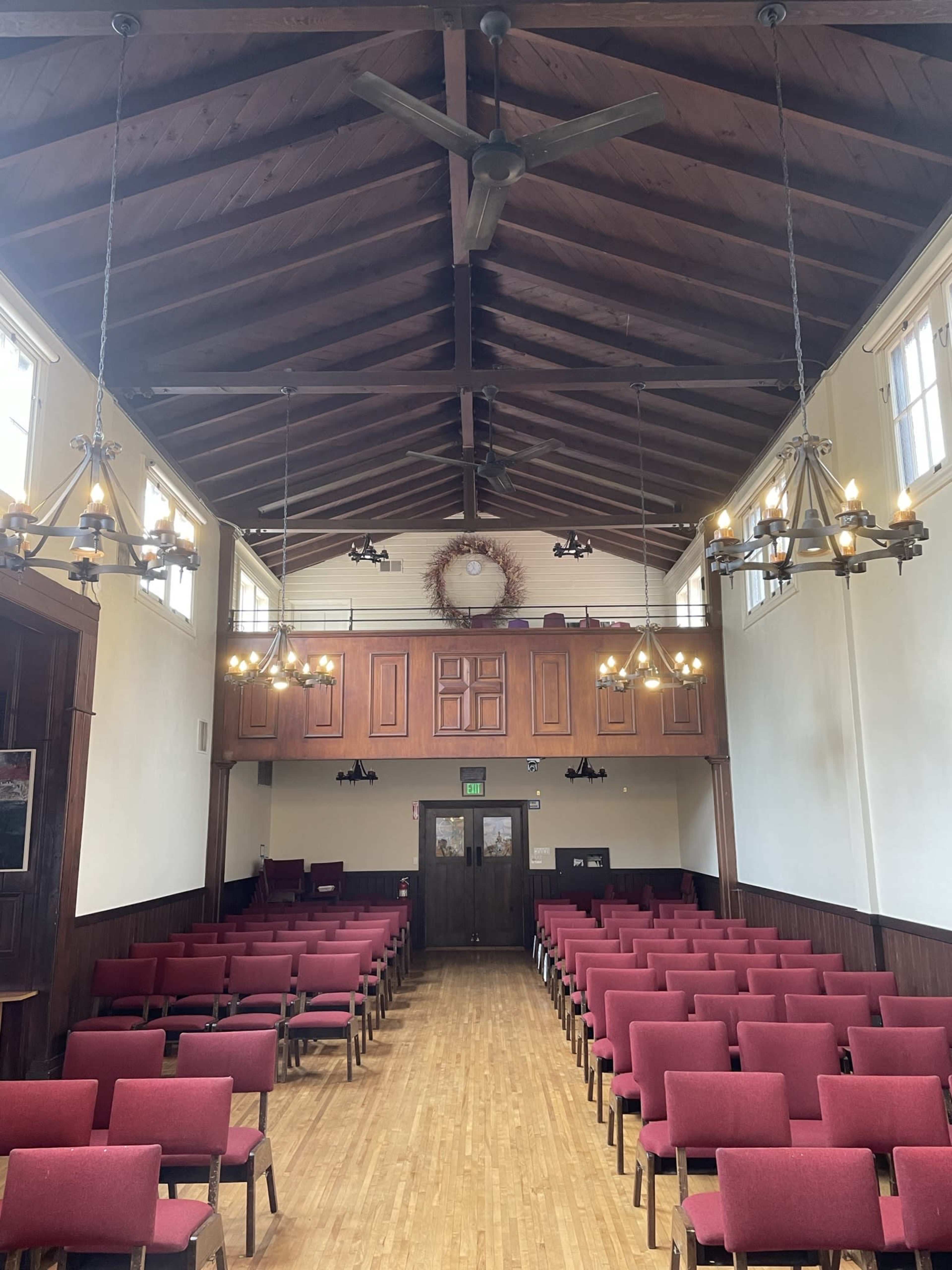 A spacious interior of a hall with rows of red chairs, wooden flooring, and a high ceiling with chandeliers.
