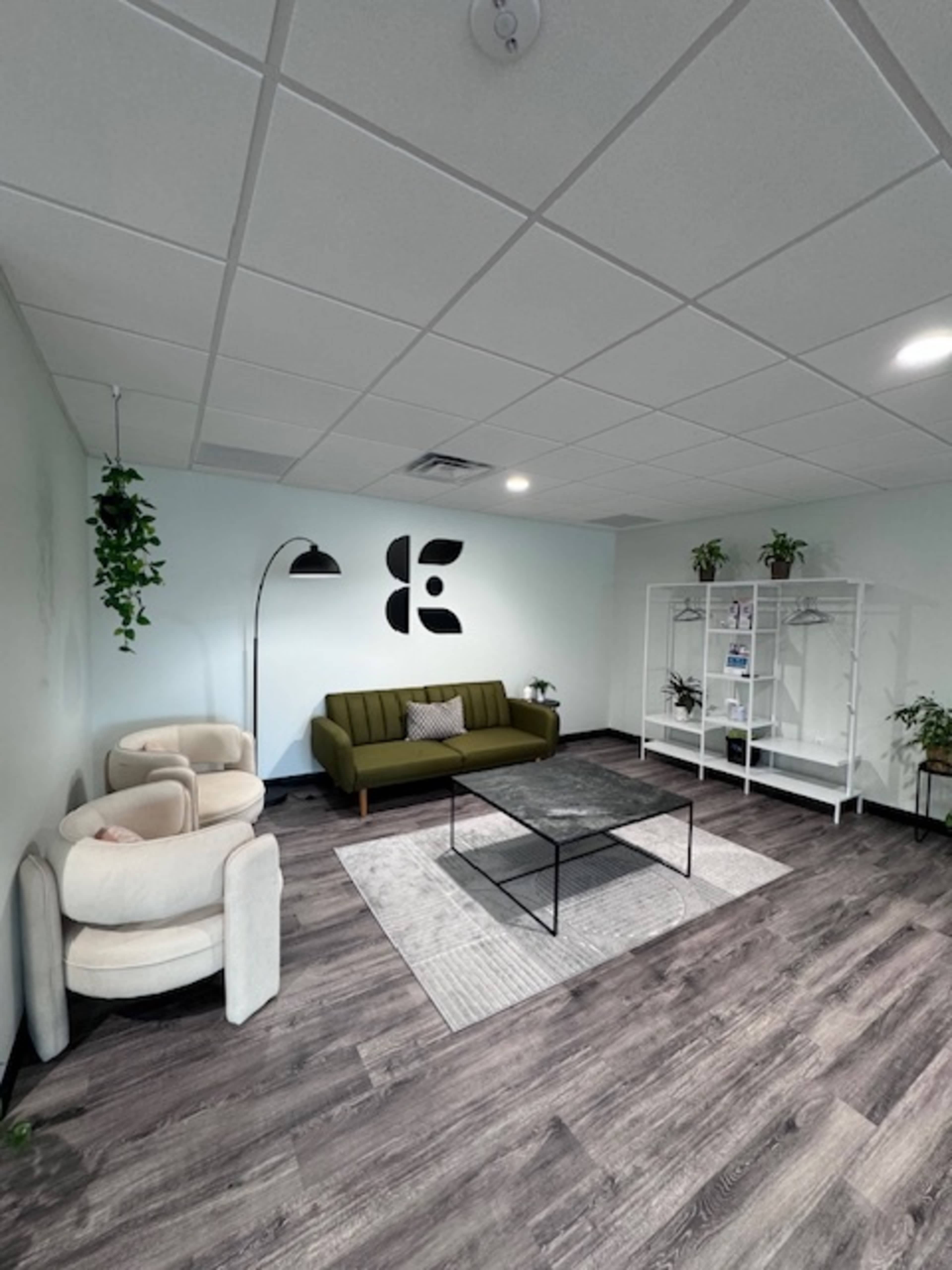 A modern office space with a green sofa, two light-colored armchairs, a black coffee table, and a bookshelf displaying plants and decor against a light-colored wall.