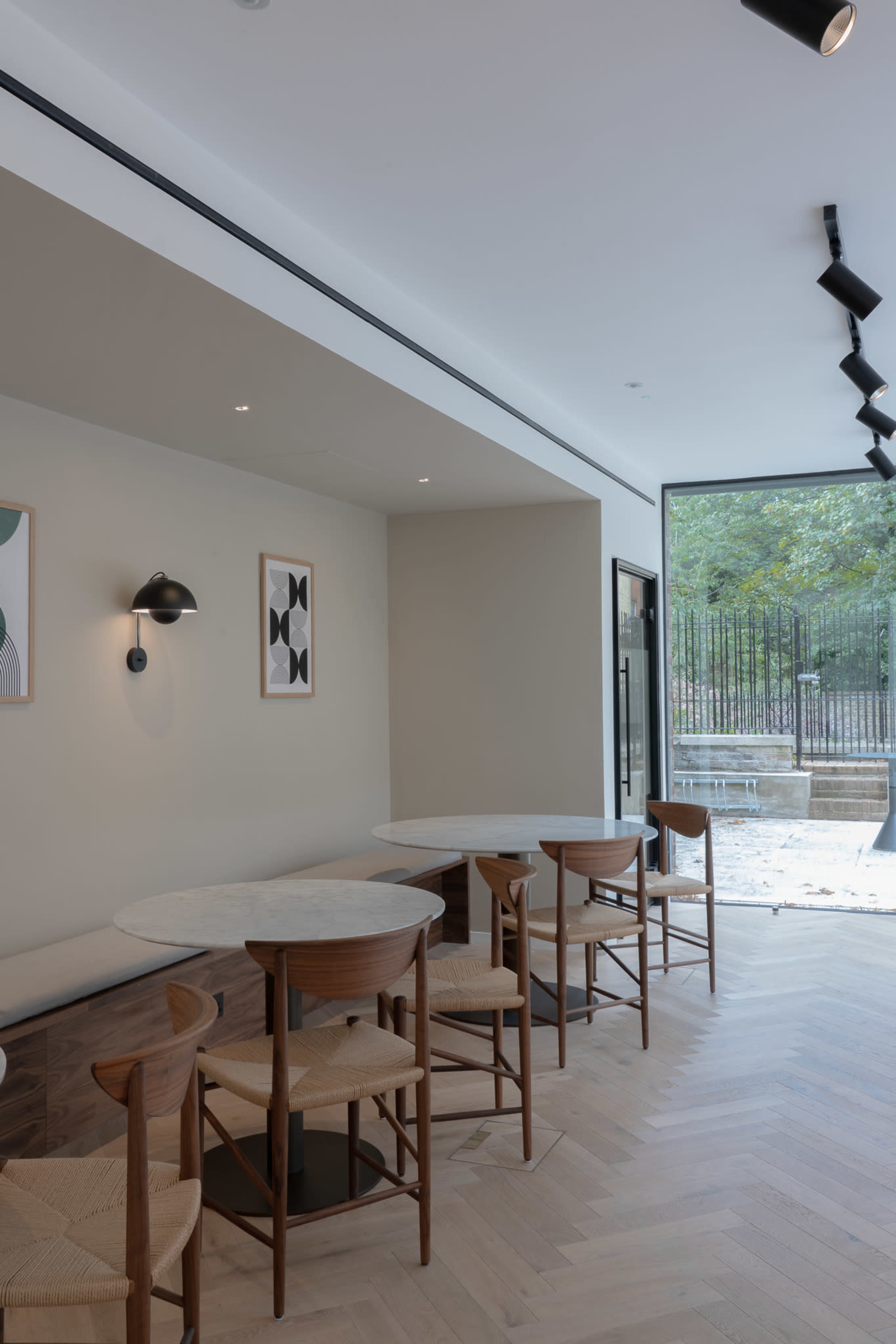 The image shows a modern café interior with light wooden flooring, round marble tables, and simple wooden chairs, featuring a large window that offers a view of an outdoor area.