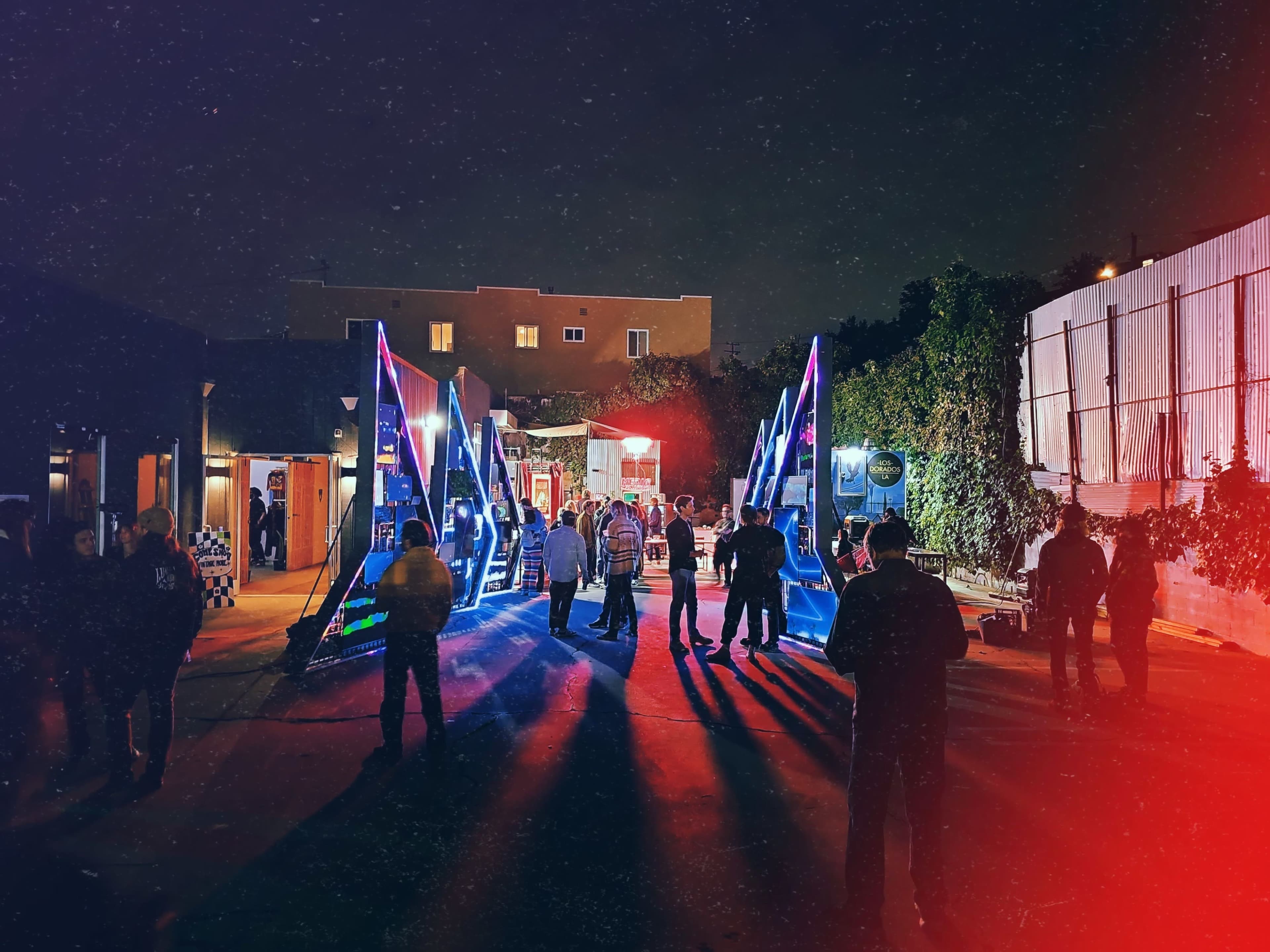 A group of people gathers in a brightly lit area with colorful lighting at night, near large, triangular structures.