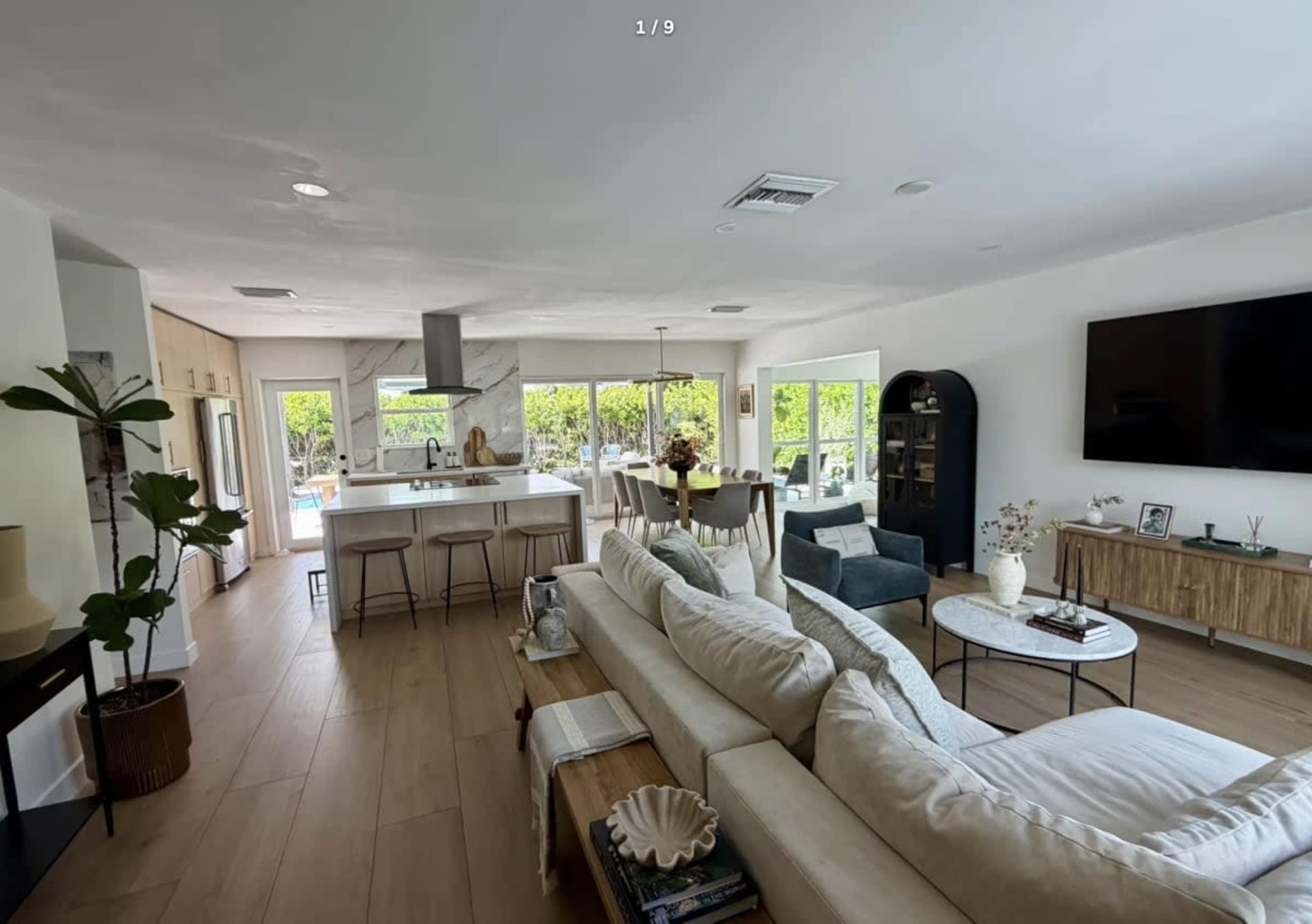 The image shows a modern open-concept living space featuring a kitchen, dining area, and seating area with large windows that provide natural light.