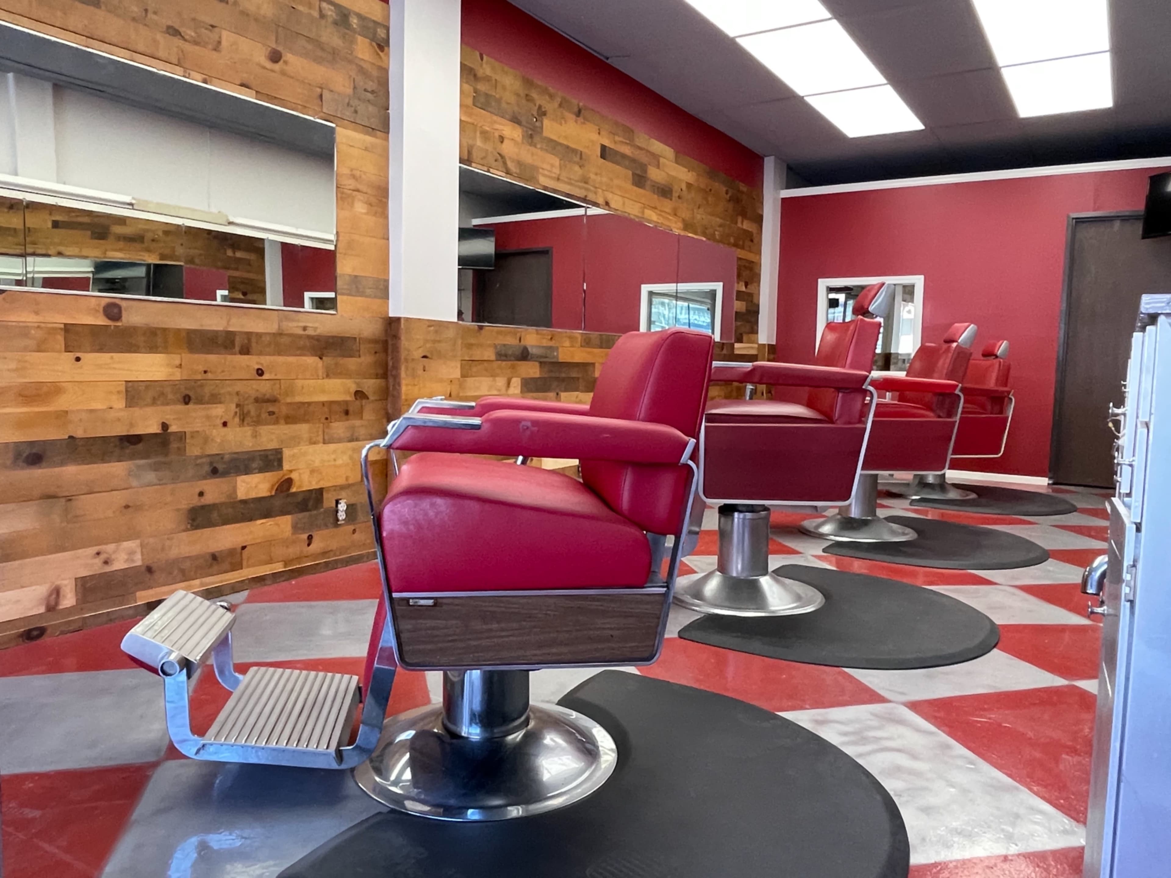 A modern barber shop interior with red and gray flooring, wooden accents on the walls, and several red barber chairs positioned in front of mirrors.