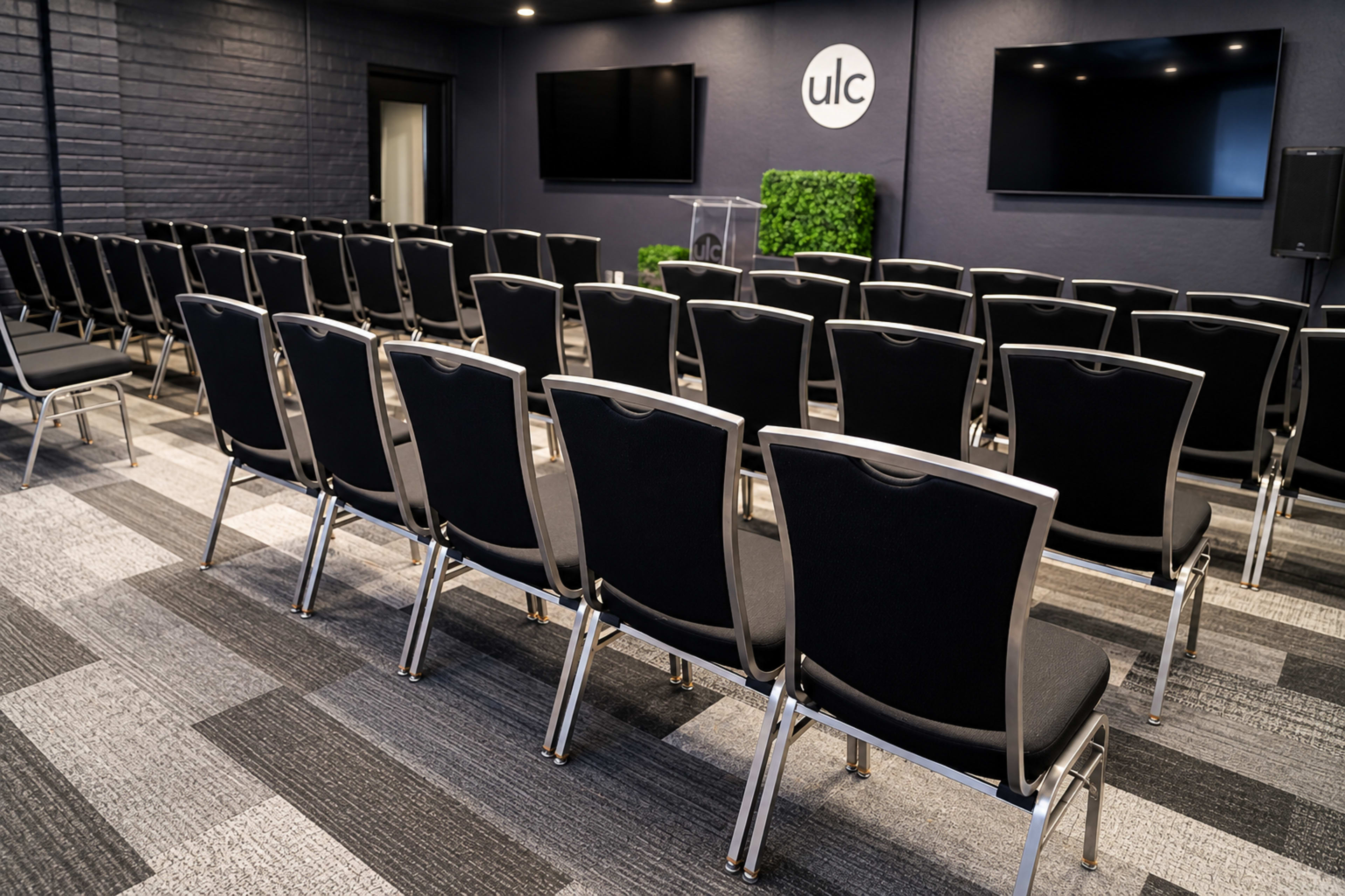 The image shows a seminar room with rows of black chairs facing a transparent podium and two wall-mounted screens.