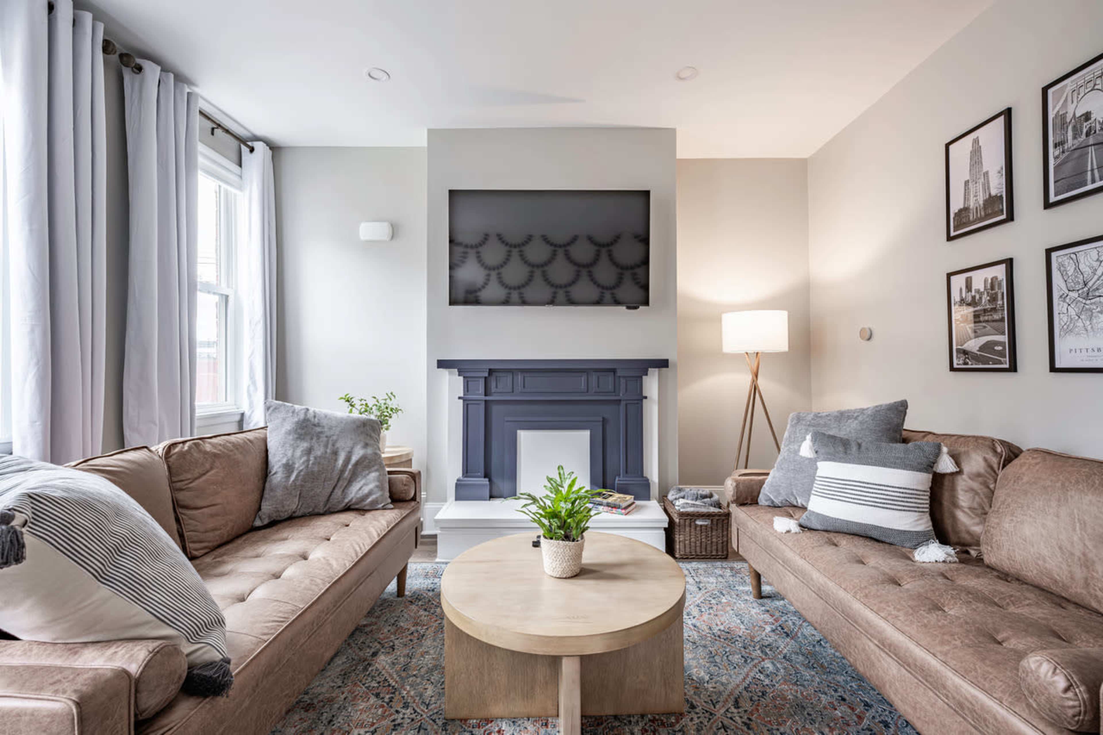 A cozy living room features two brown sofas facing a circular wooden coffee table, a blue fireplace with a TV above, and light gray curtains.
