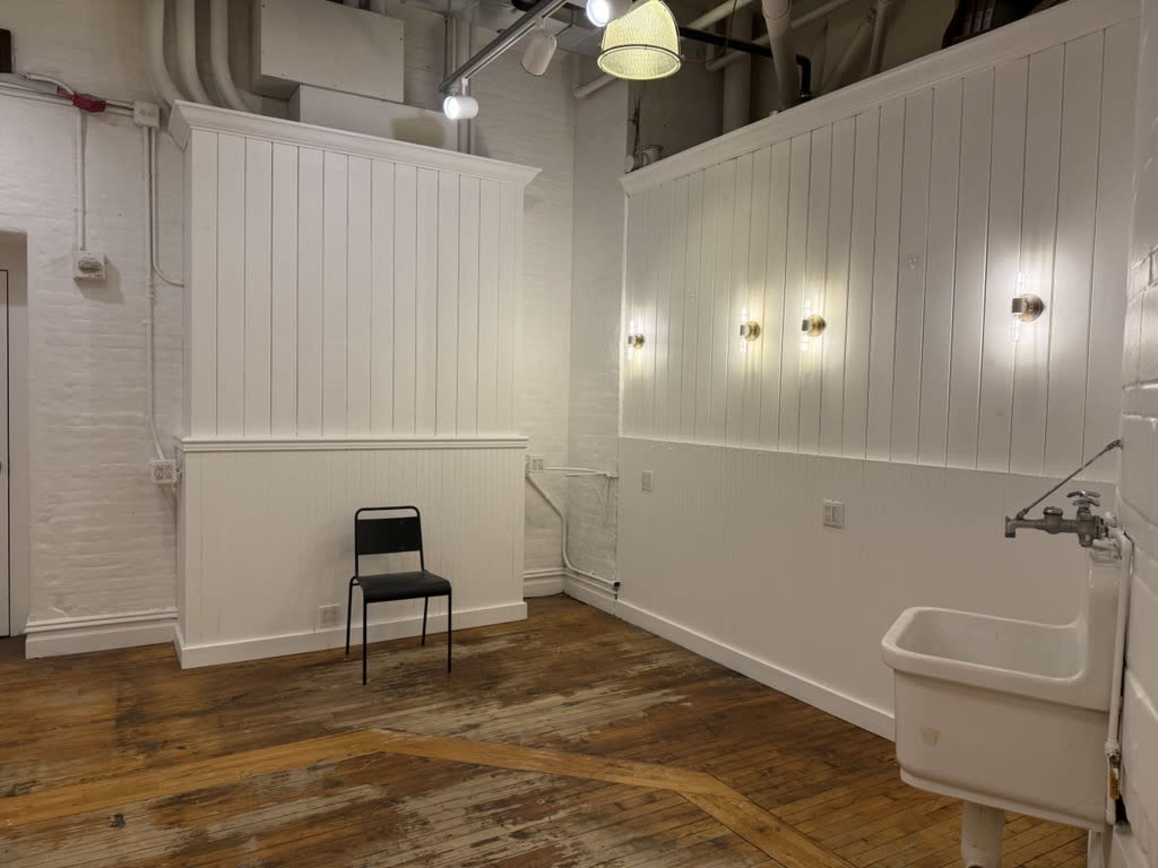 The image shows a sparsely furnished room with white wooden walls, a black chair, a wall-mounted sink, and several light fixtures illuminating the space.